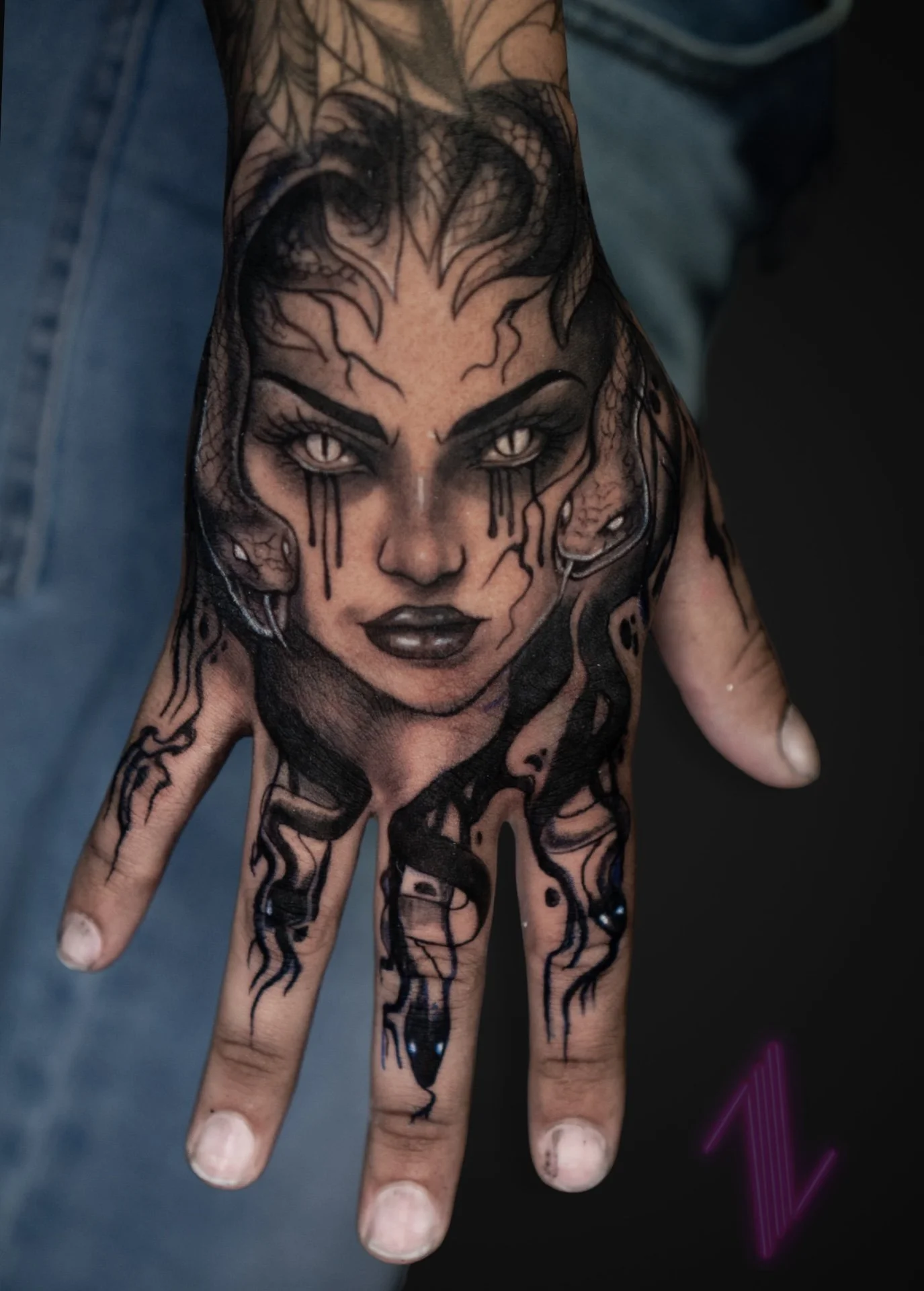 Tattoo of a woman's face with dark makeup, piercing eyes, and ink dripping down her face, on a person's hand.