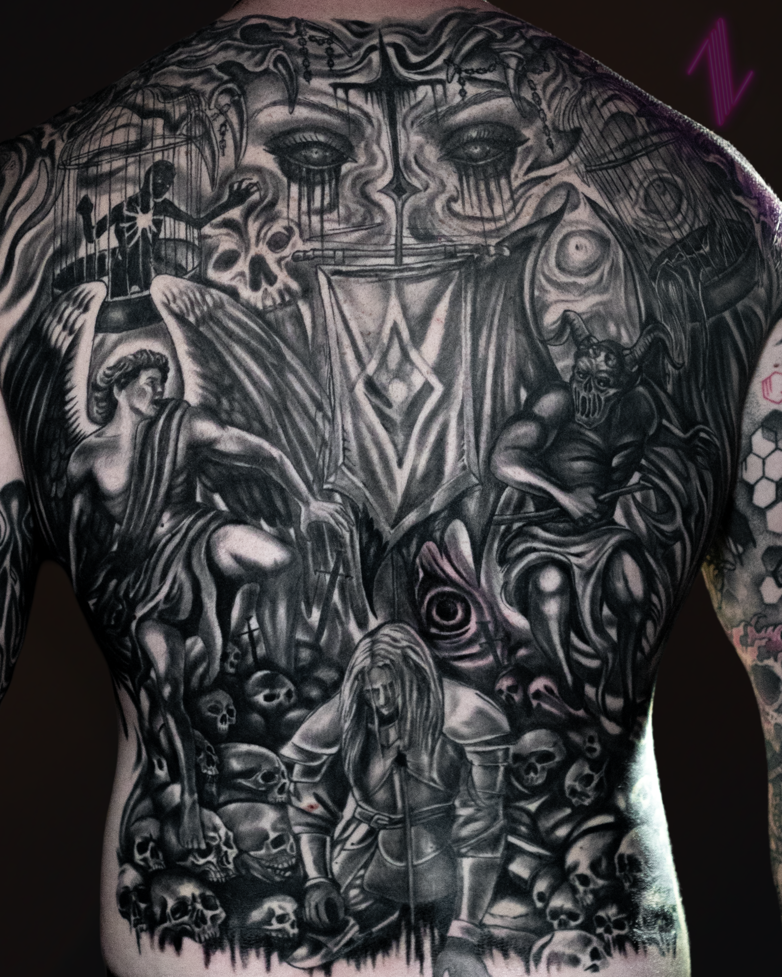 Black and gray tattoo covering a person's back, featuring a large face with dripping eyes at the top, surrounded by various detailed figures including angels, demons, skulls, and mystical symbols.