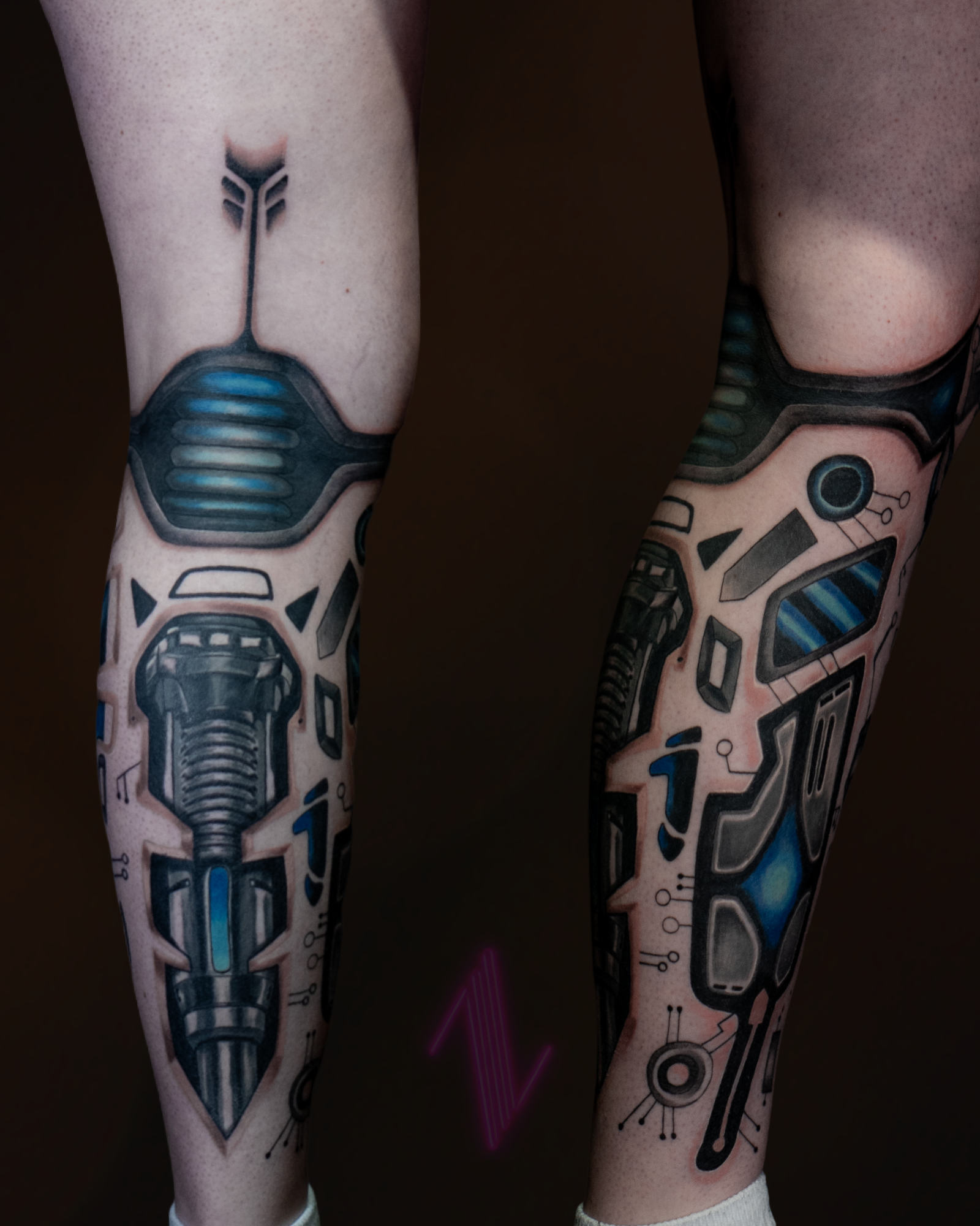 Close-up of two human legs with futuristic robotic tattoos in black and blue, covering from the thighs to the calves.