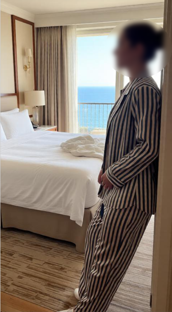 A person in striped pajamas standing in a hotel room near a bed with white linens and a towel folded on it, with a large window showing the ocean in the background.