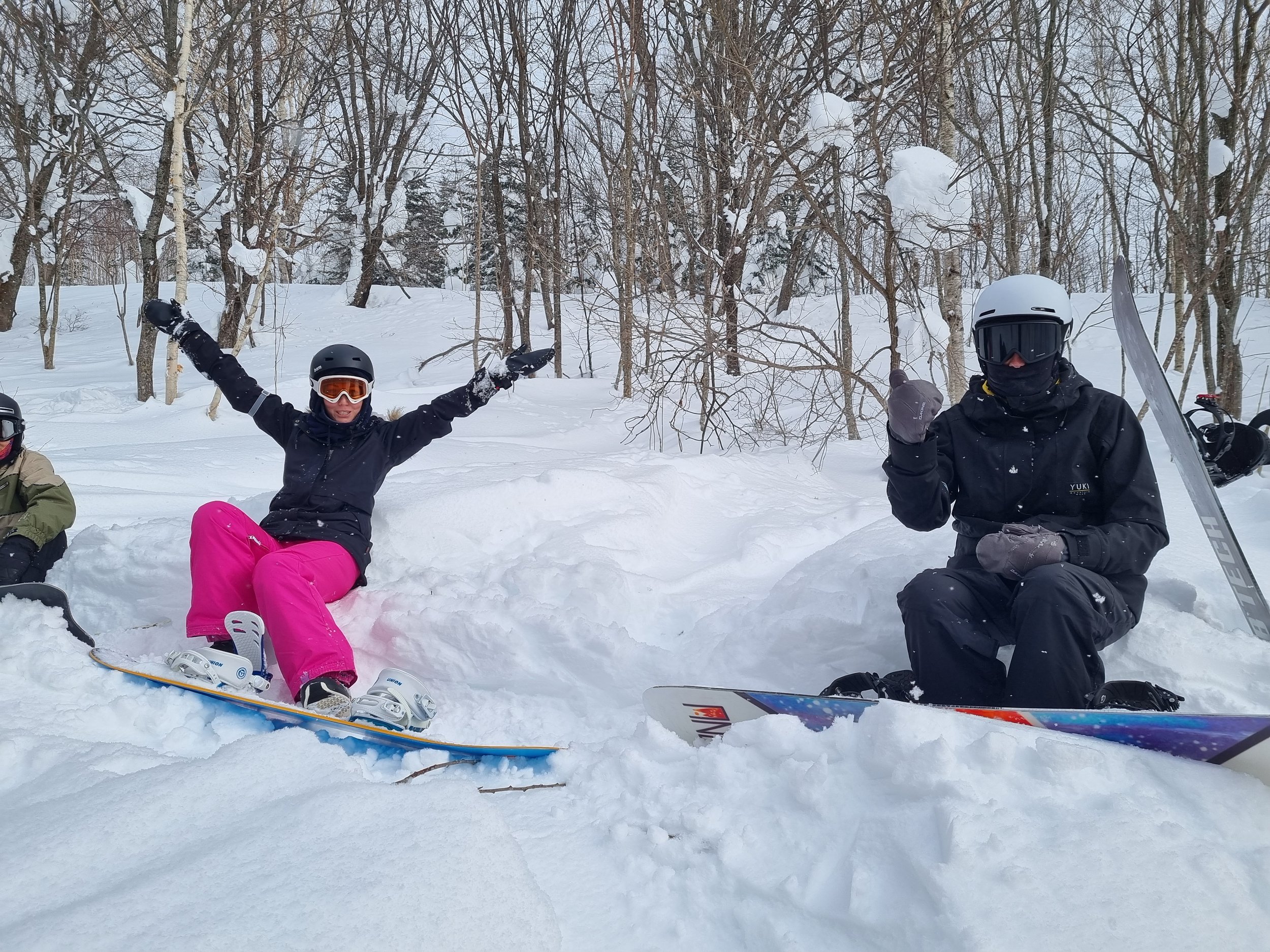 Two people in winter gear sitting on snow with trees in the background, one woman with pink pants and goggles raising her arms, and one person wearing a helmet and goggles giving a thumbs-up, both sitting next to skis.