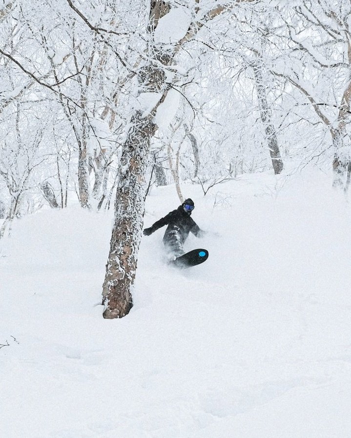 Snowboarder in snowy forest, wearing dark clothing and goggles, navigating through snow-covered trees.