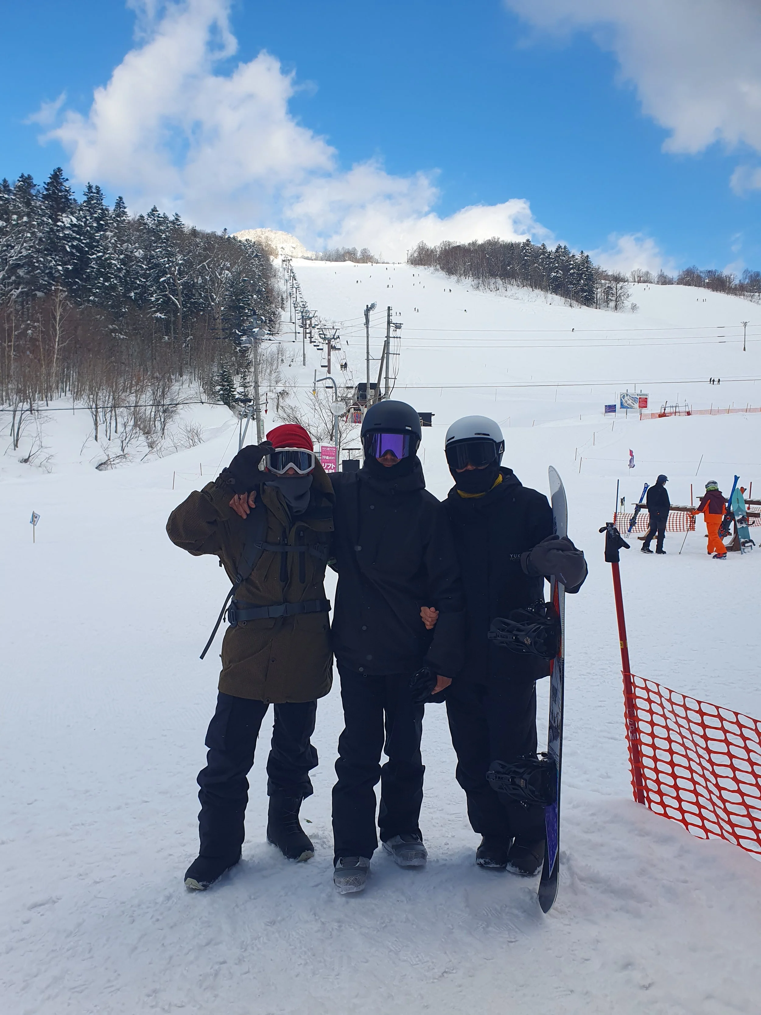 Three snowboarders in winter gear posing on a snowy slope with a ski lift and forest in the background in Hokkaido Japan.
