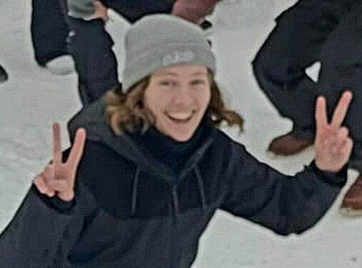 A person in a black jacket and gray beanie smiling, making peace signs with both hands in a snowy setting.