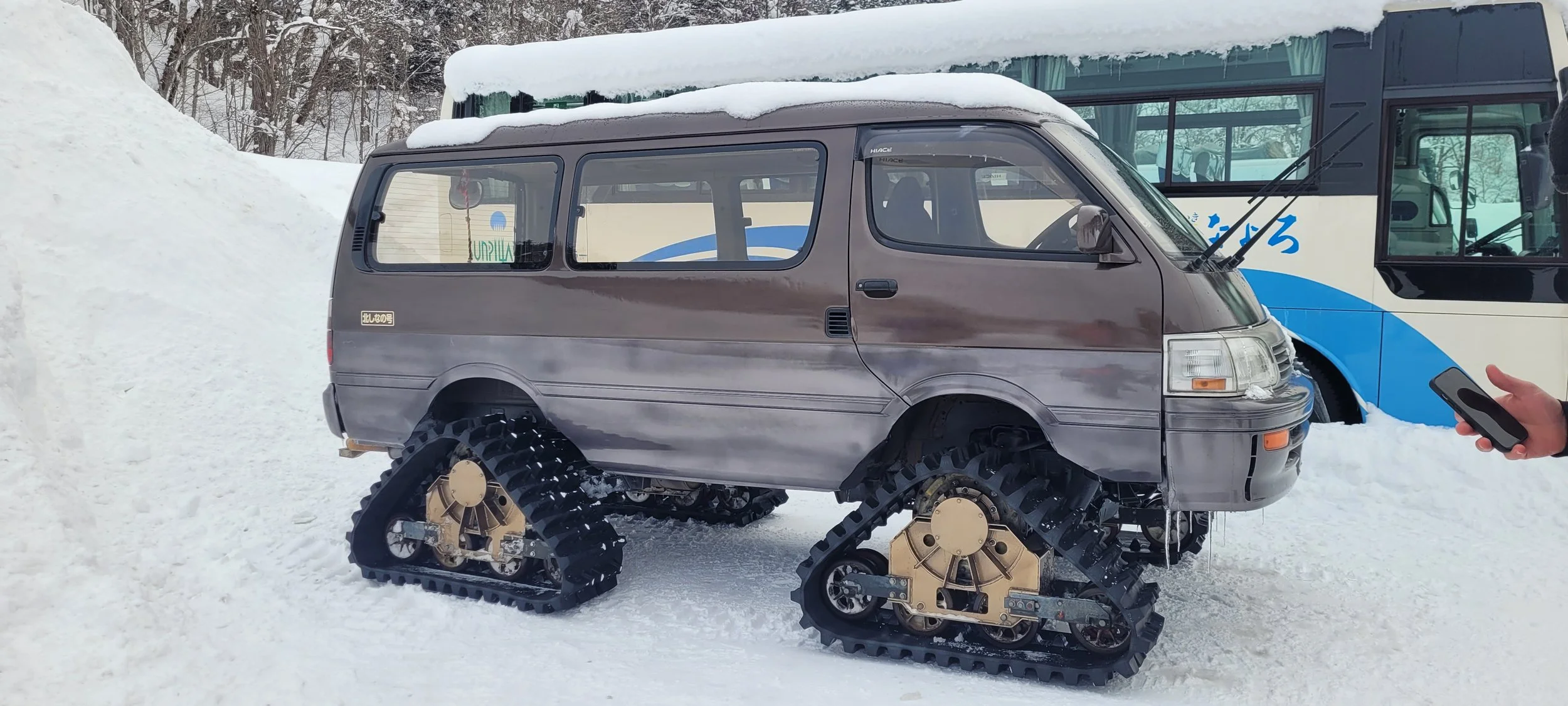 A van equipped with snow tracks instead of regular wheels, parked on a snowy surface beside a bus in Hokkaido Japan.
