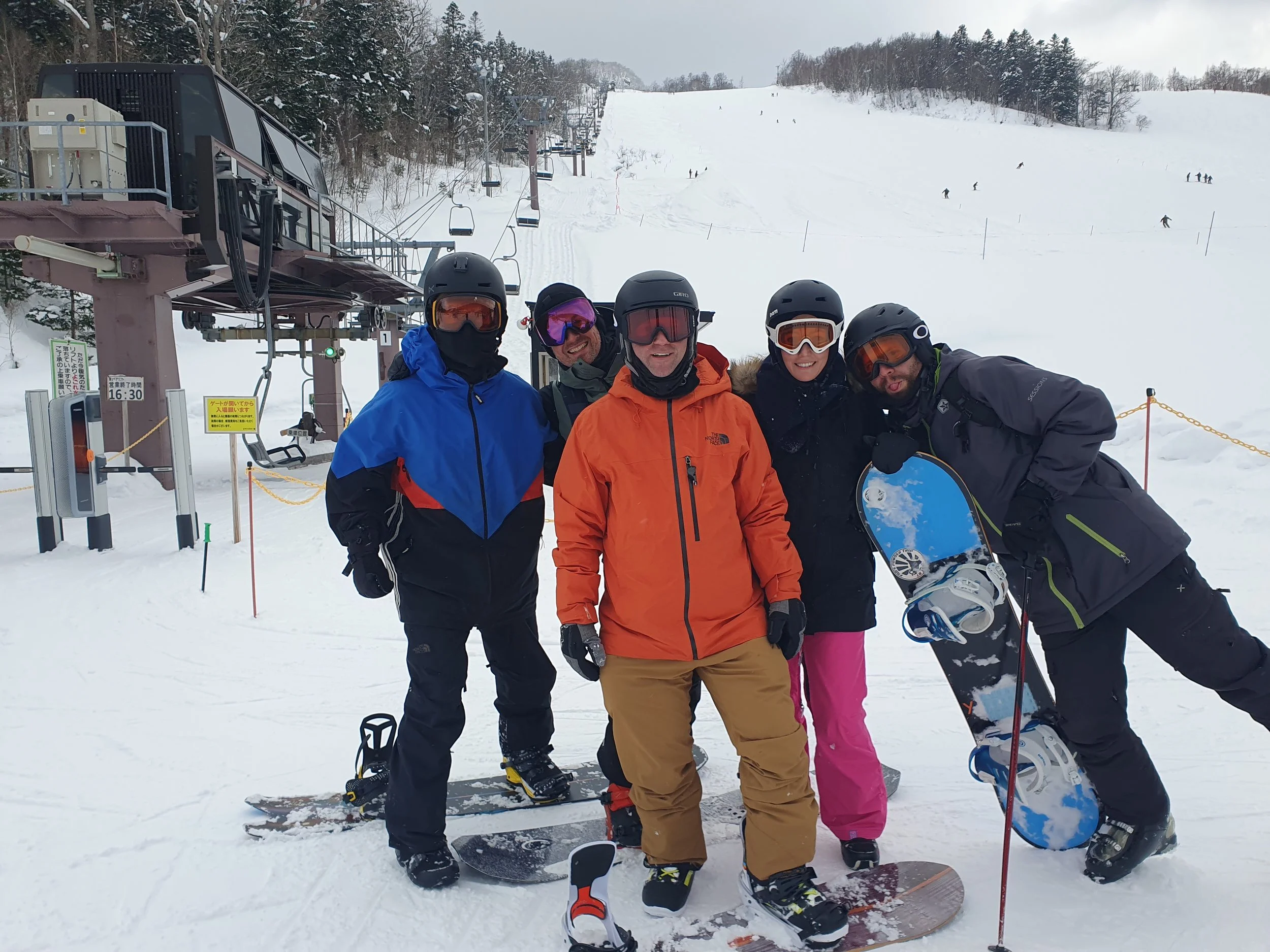 Group of snowboarders posing at a ski resort with a chairlift and snowy slope in the background in Hokkaido Japan.