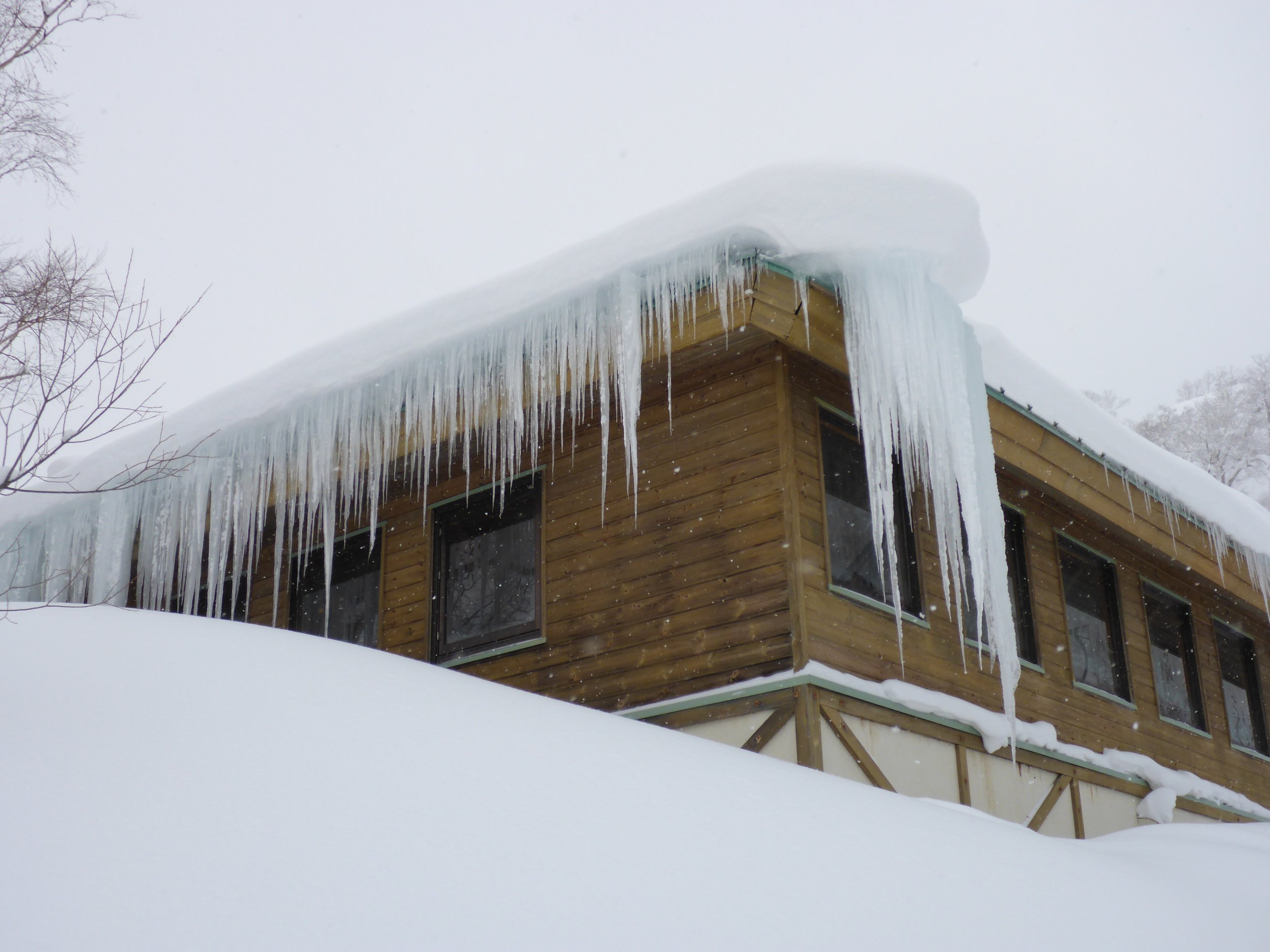 A wooden house with large icicles hanging from the snow-covered roof, surrounded by deep snow.
