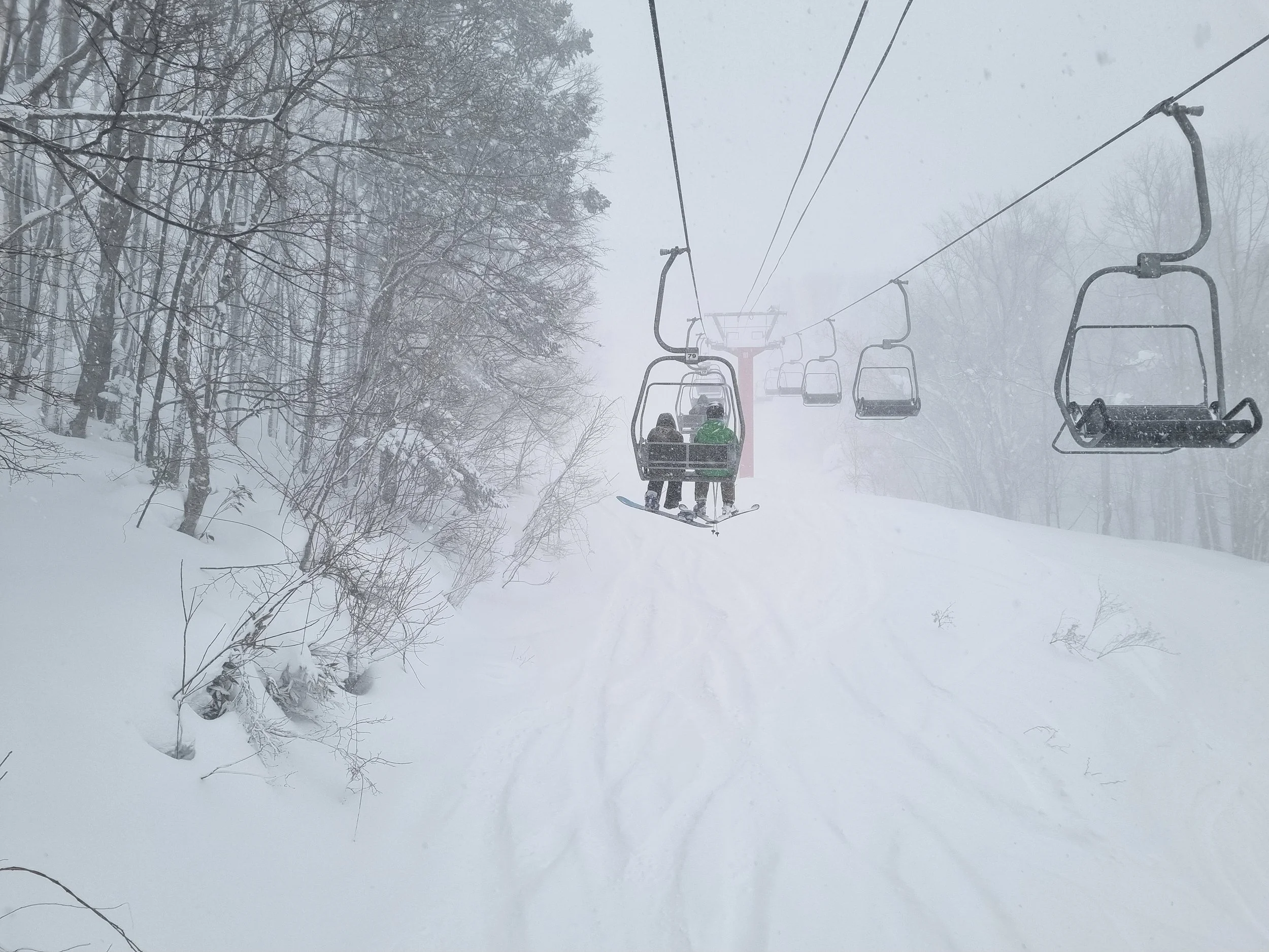 Ski lift chairs traveling up a snowy mountain with two skiers on one of the chairs, surrounded by snow-covered trees and falling snow.