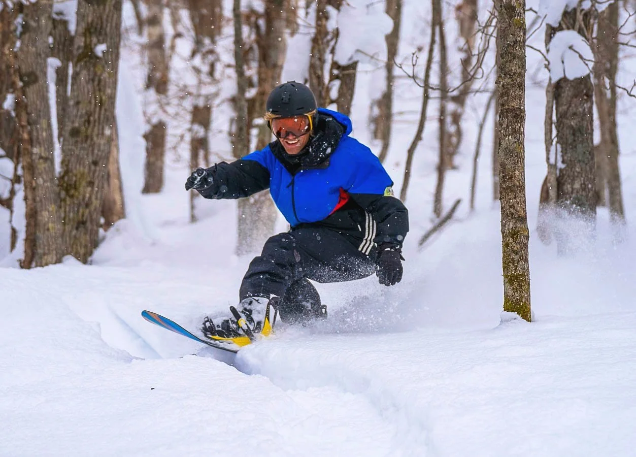 A person snowboarding through a snowy forest with trees and snow-covered ground.