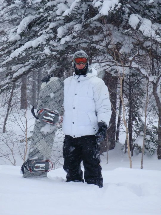 Person in winter gear holding a snowboard while standing in snow-covered forest