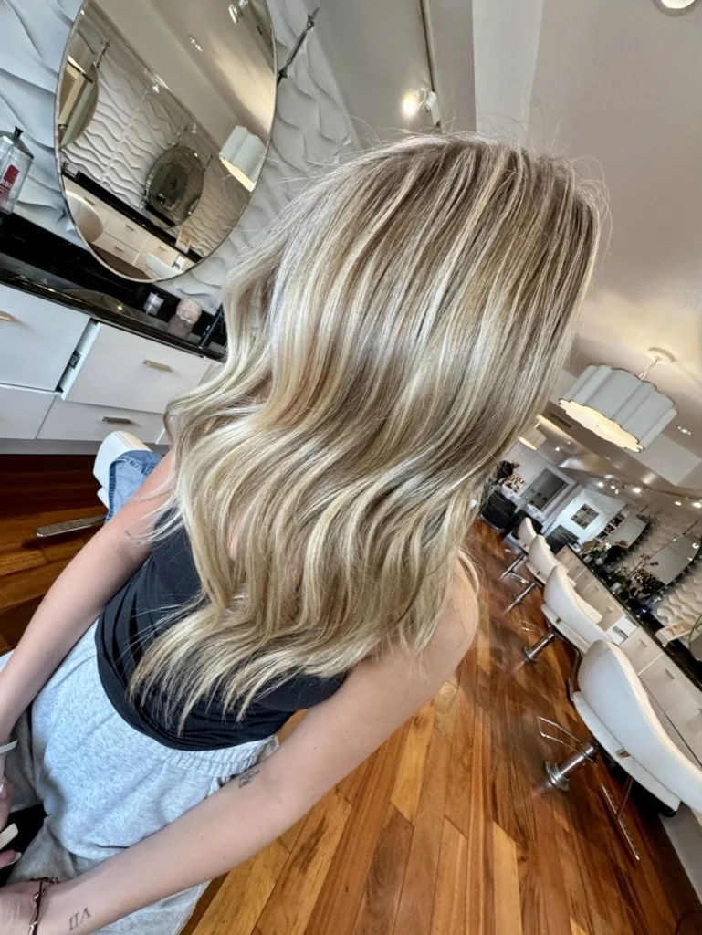 A woman with wavy blonde hair sitting in a salon with modern decor, white chairs, and a wooden floor.