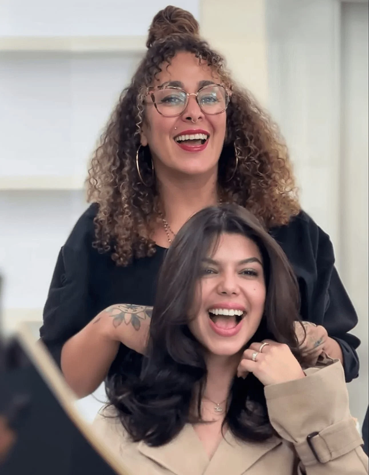 Two women smiling and laughing, one with curly hair and glasses, and the other with dark hair and tattoos, sitting close together in a bright room.
