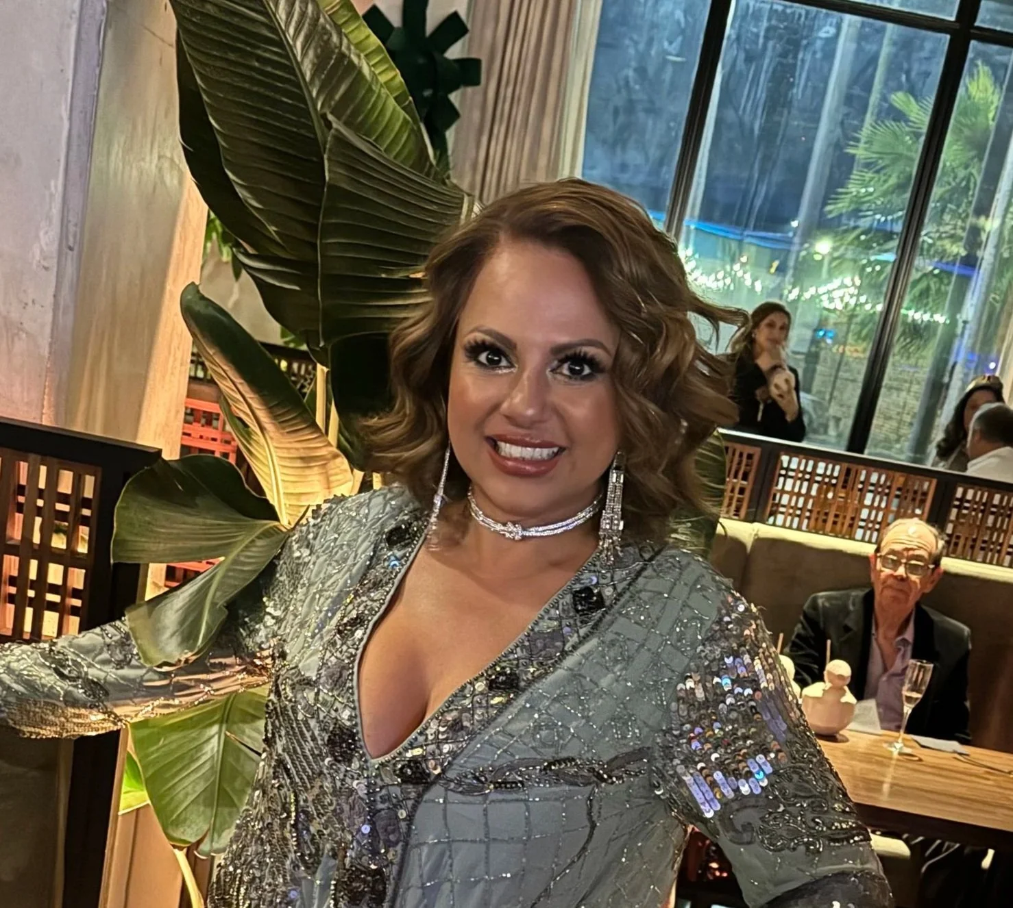 A woman with wavy, shoulder-length brown hair smiling at the camera, dressed in a sparkly, sequined silver dress, wearing large earrings and a necklace, in an indoor setting with large green leafy plants and other people in the background.