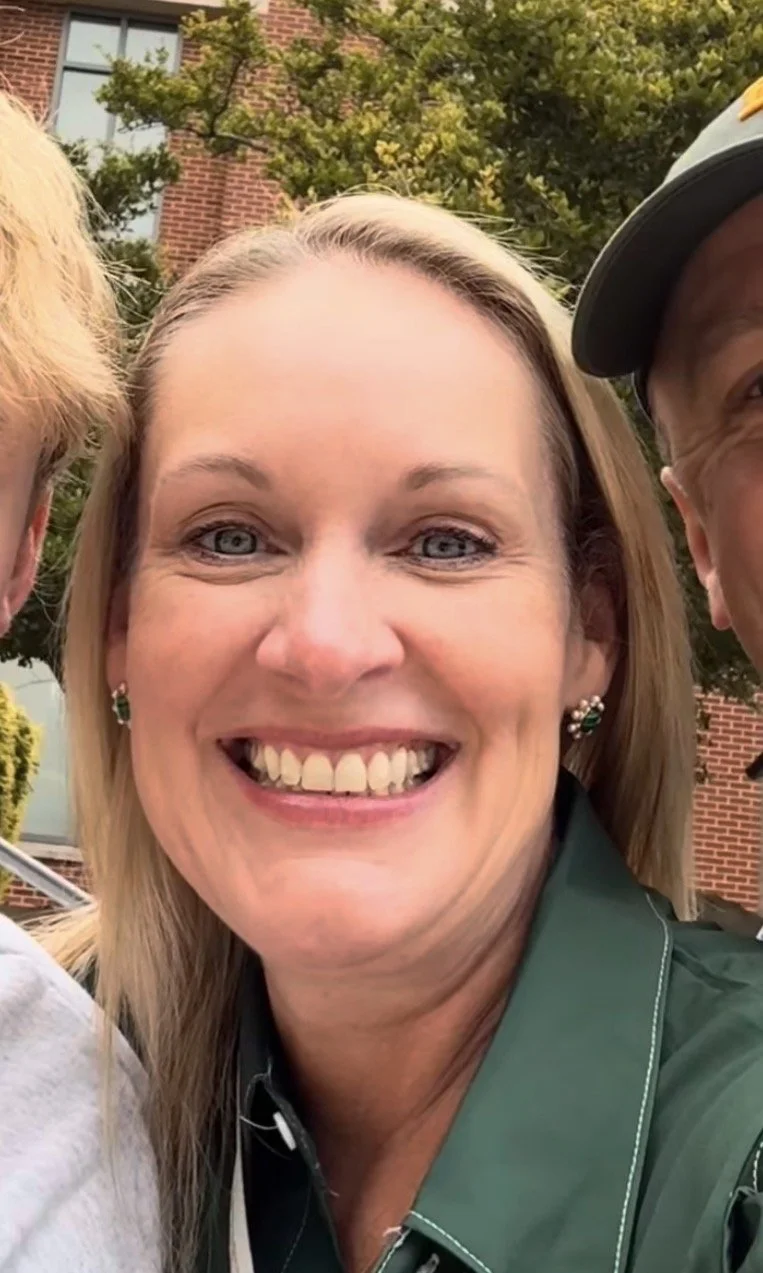 Close-up of a smiling woman with blonde hair, blue eyes, and earrings, outdoors with trees and a brick building in the background.