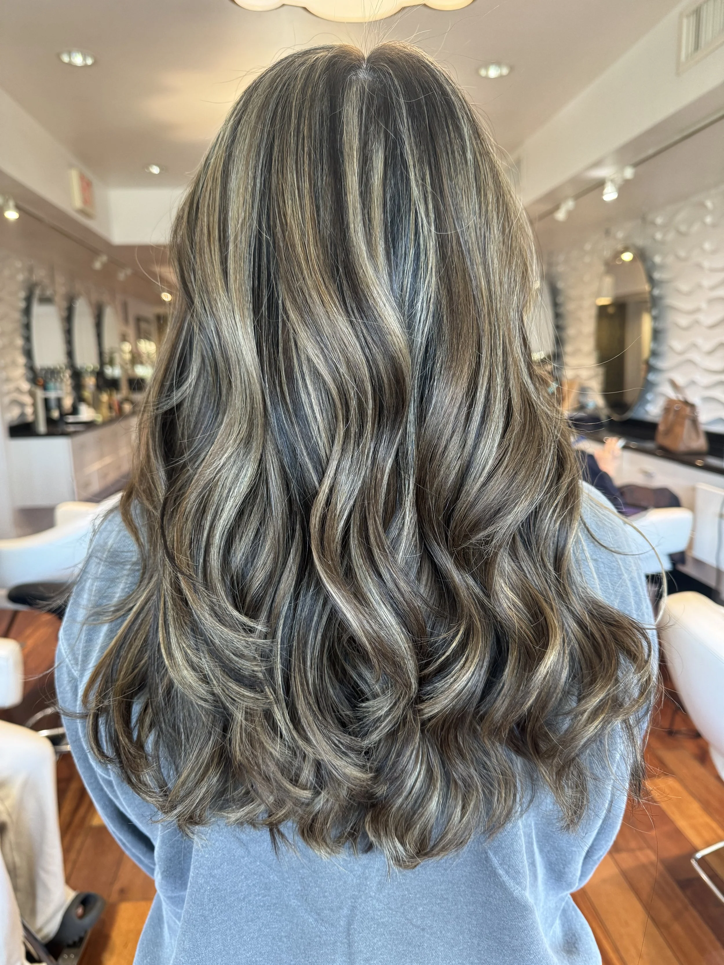 A person with long, wavy, blonde hair sitting on a stool in a salon. The background includes salon mirrors, lights, and fixtures.