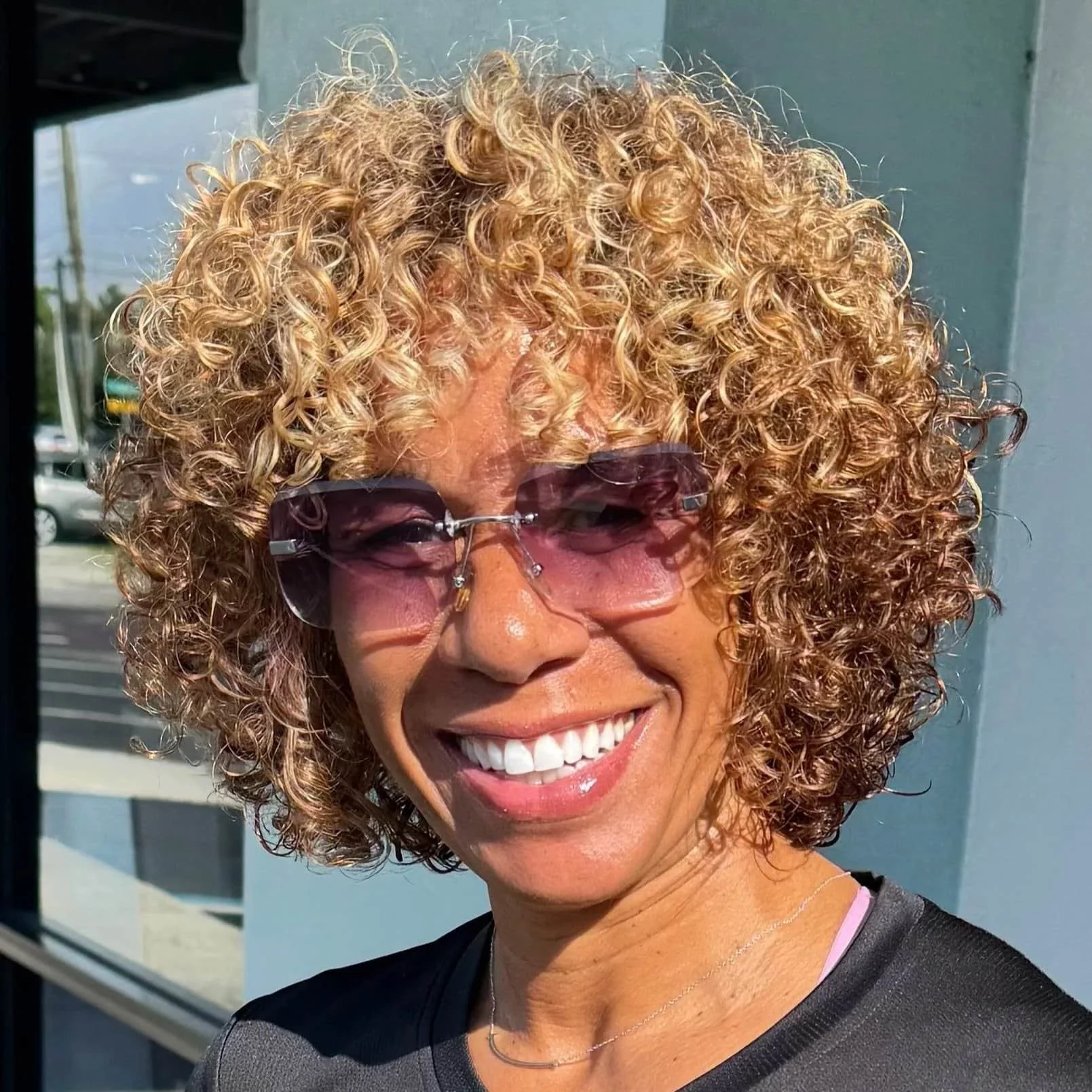 A woman with light brown skin and curly blonde hair wearing sunglasses and a black top, smiling outdoors.