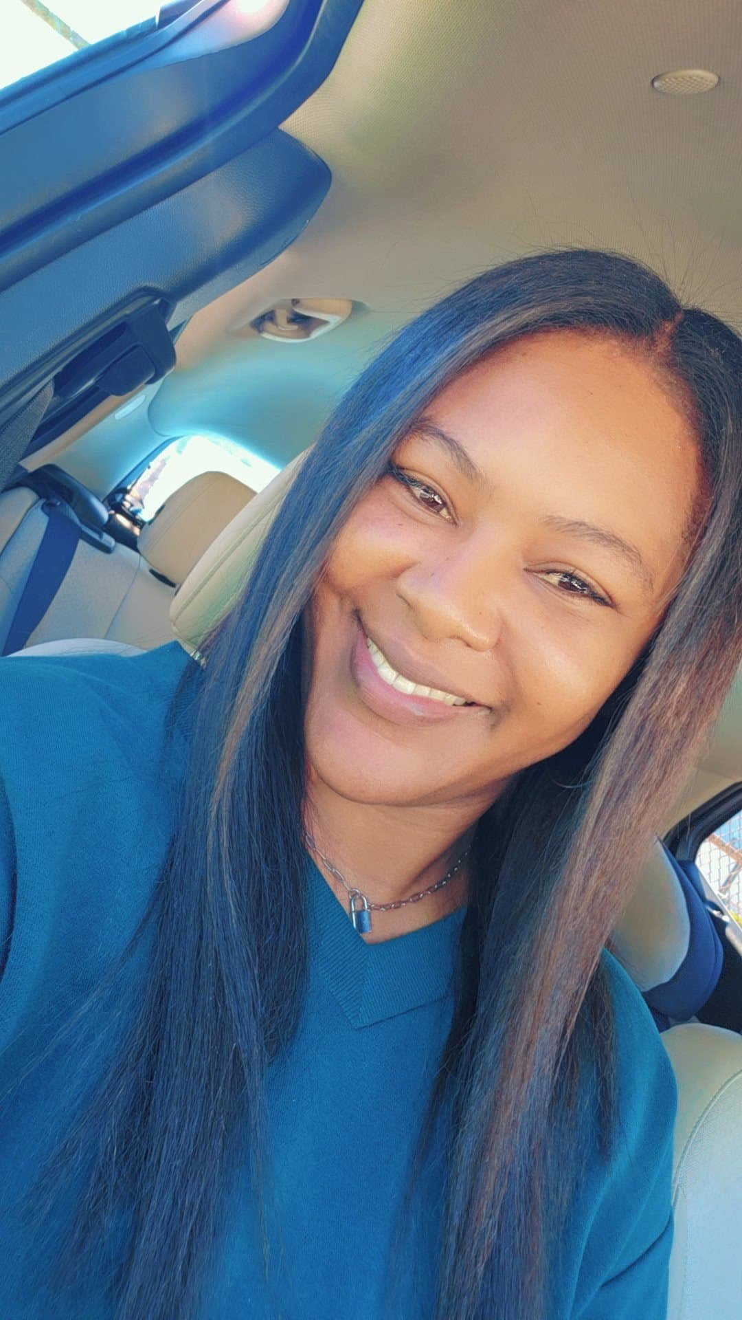 A woman smiling and sitting in a car, with long dark hair, wearing a blue shirt and a silver chain necklace.