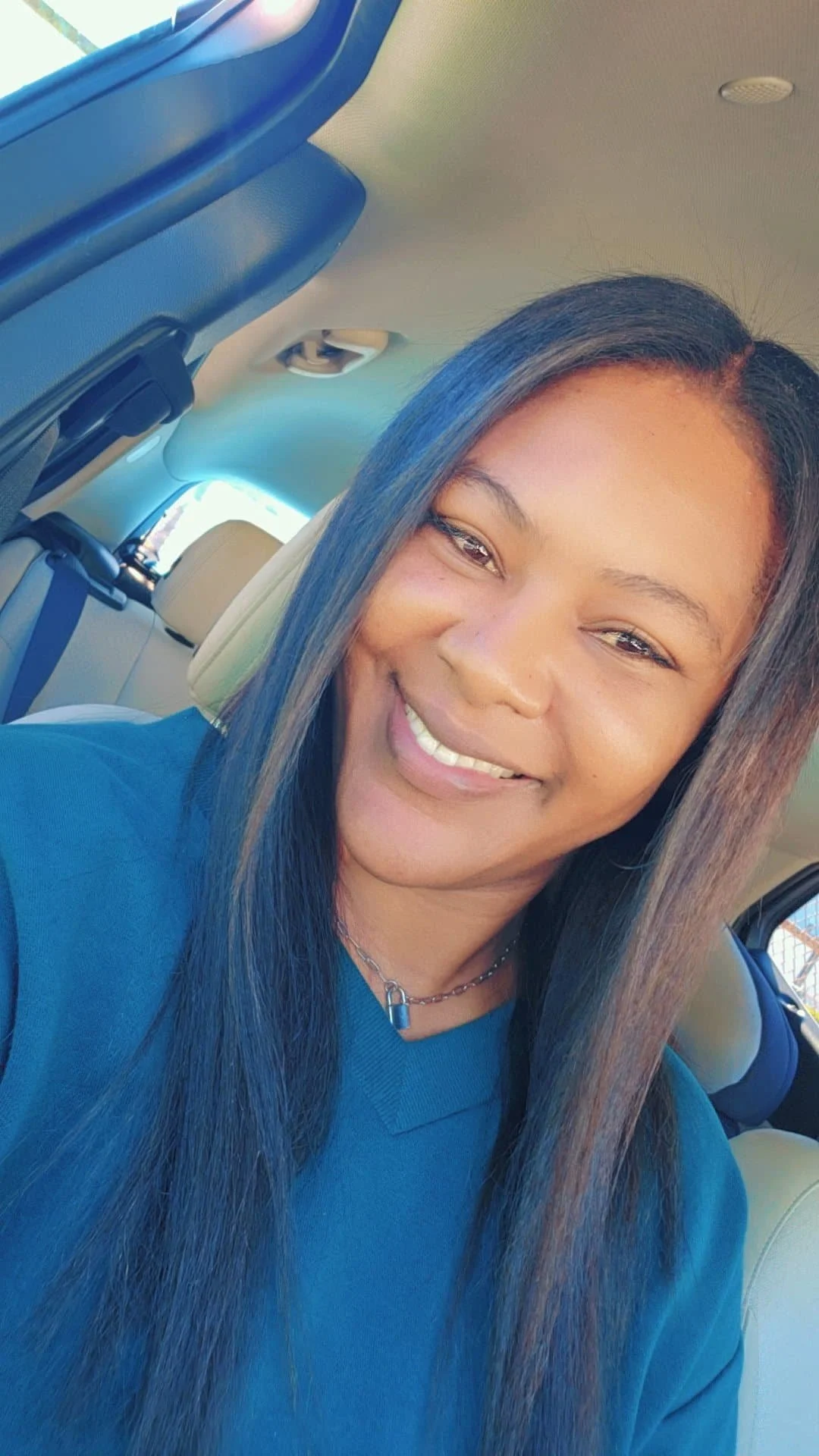 A woman smiling and sitting in a car, with long dark hair, wearing a blue shirt and a silver chain necklace.