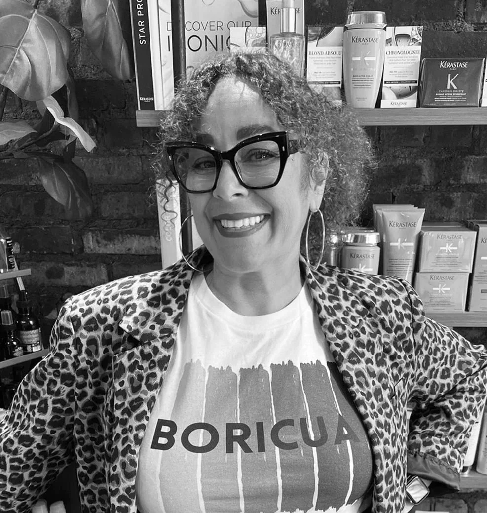 A woman with curly hair, glasses, and hoop earrings, wearing a leopard-print jacket and a t-shirt that says "Boricua," smiling and standing in front of shelves with hair care products.