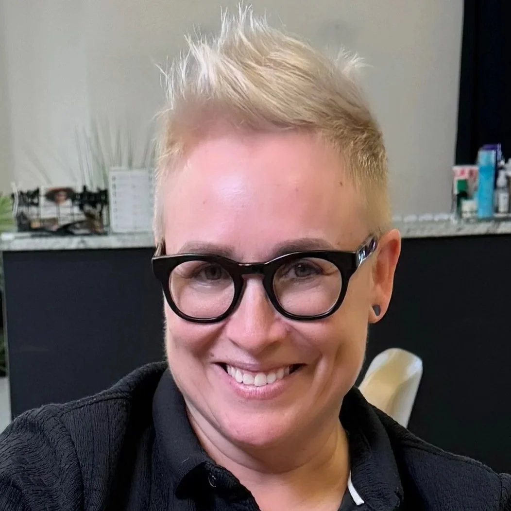 Close-up of a smiling woman with short blond hair, black glasses, and a black shirt, standing indoors with a black counter and kitchen items in the background.