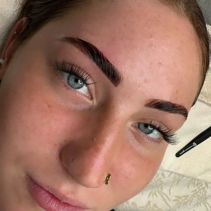 Close-up of a woman with light skin, blue eyes, dark eyebrows, and long eyelashes, lying down with her head tilted slightly. A marker or pen is visible near her face.