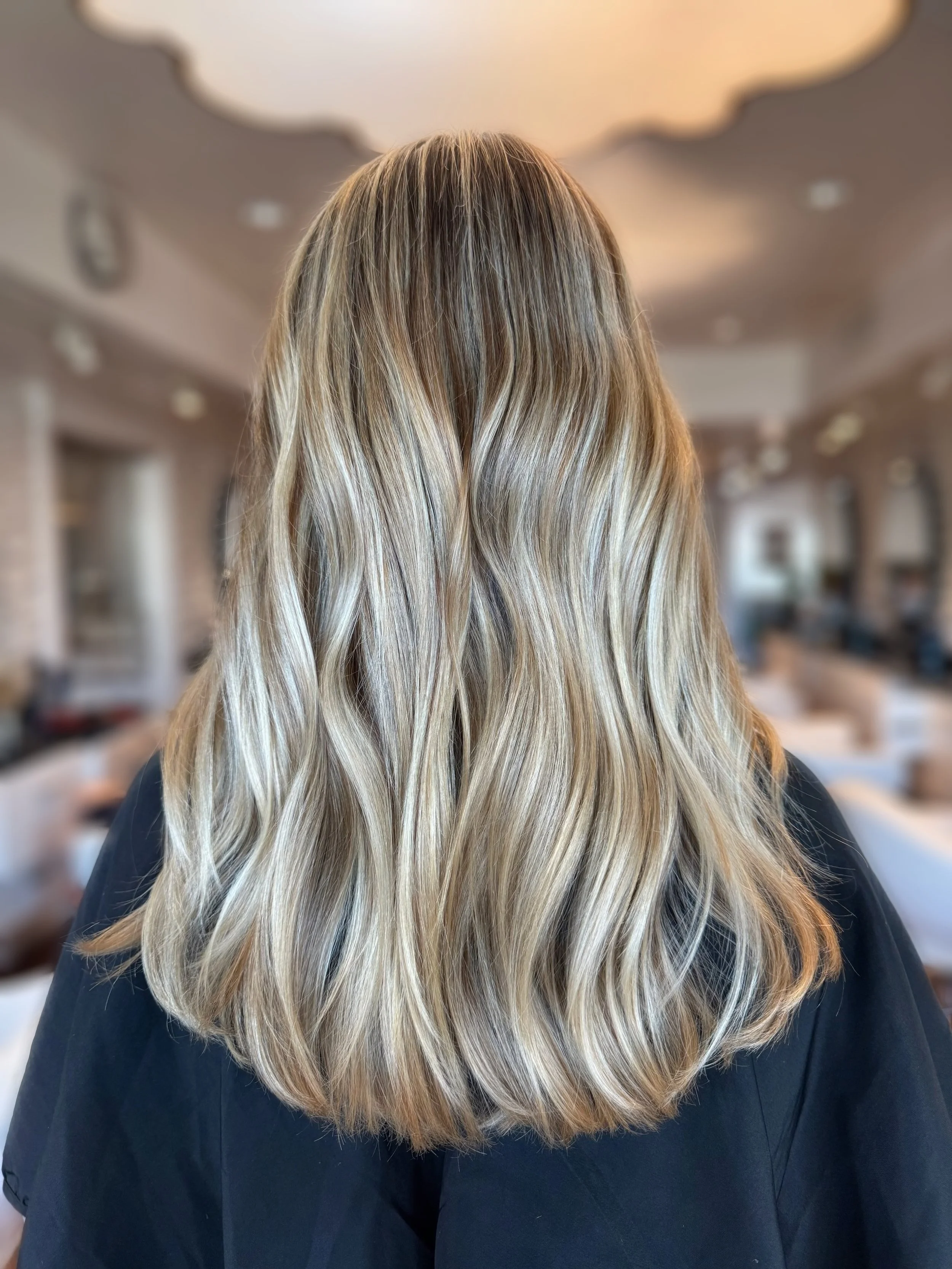 Back view of a woman with long, wavy blonde hair in a salon.