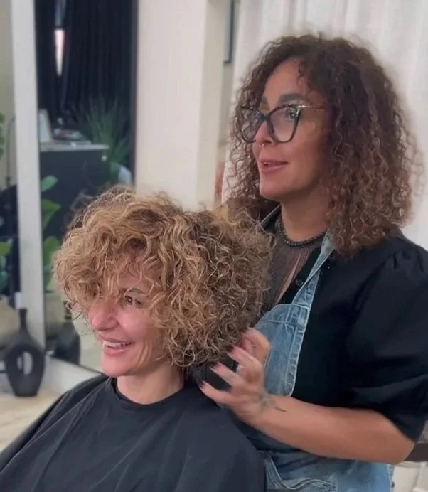 A hairstylist with curly hair and glasses styling a woman's curly hair in a salon.