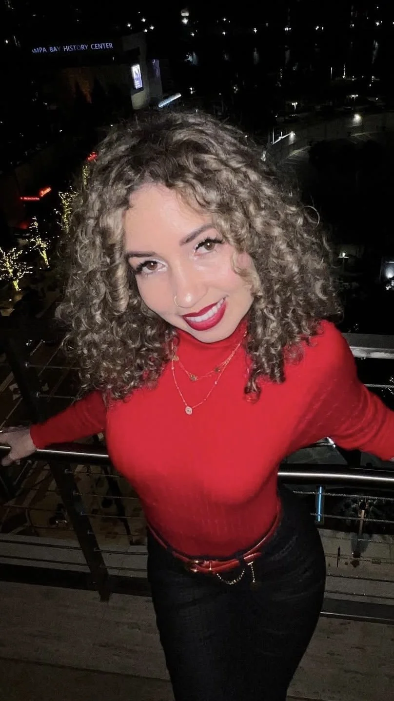 A woman with curly blonde hair wearing a red top and black pants. She is smiling and standing outdoors at night near a railing with a cityscape in the background, including lit trees and buildings.
