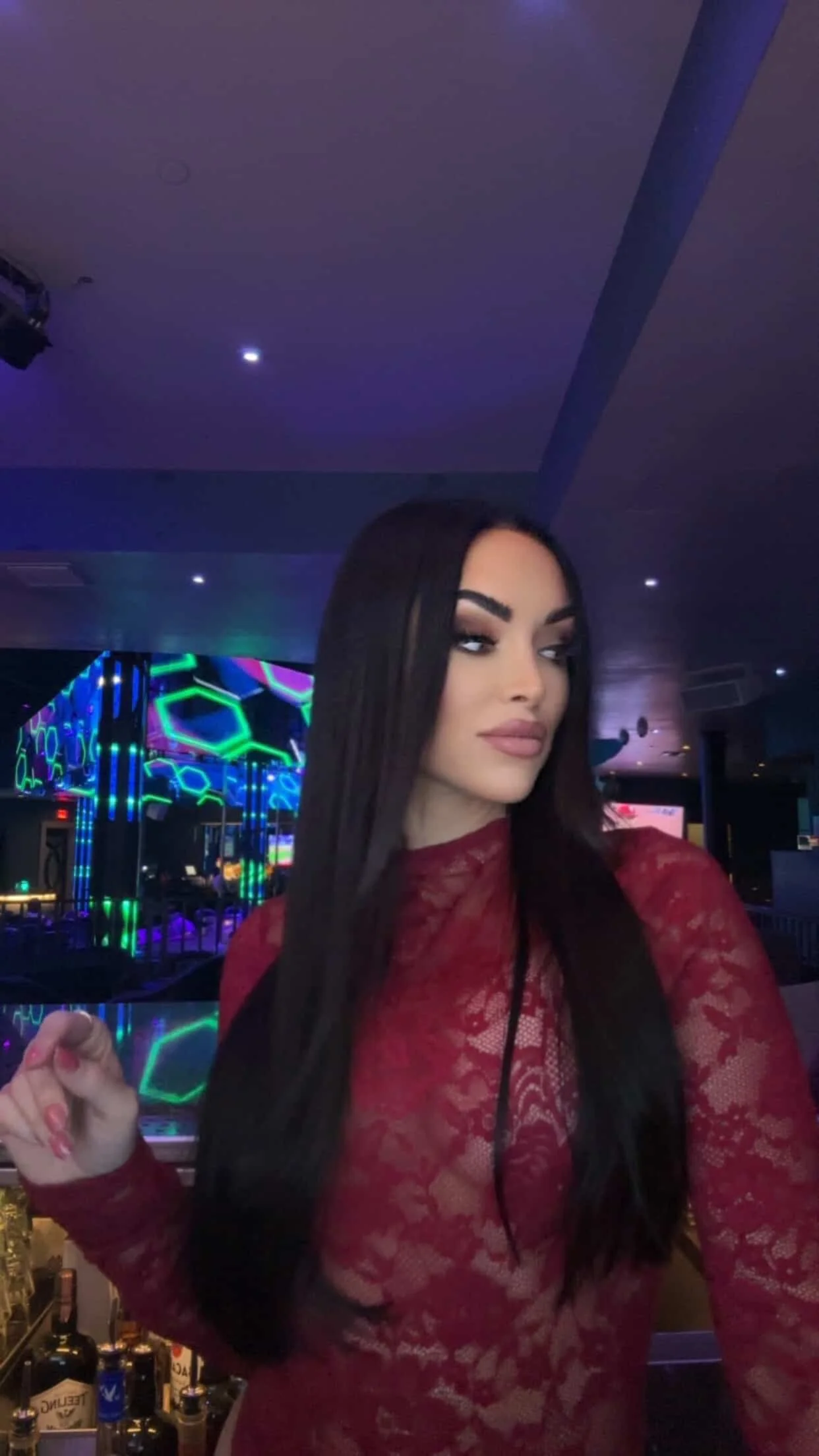 A woman with long dark hair, wearing a red lace dress, is in a dimly lit area with colorful neon lights in the background.