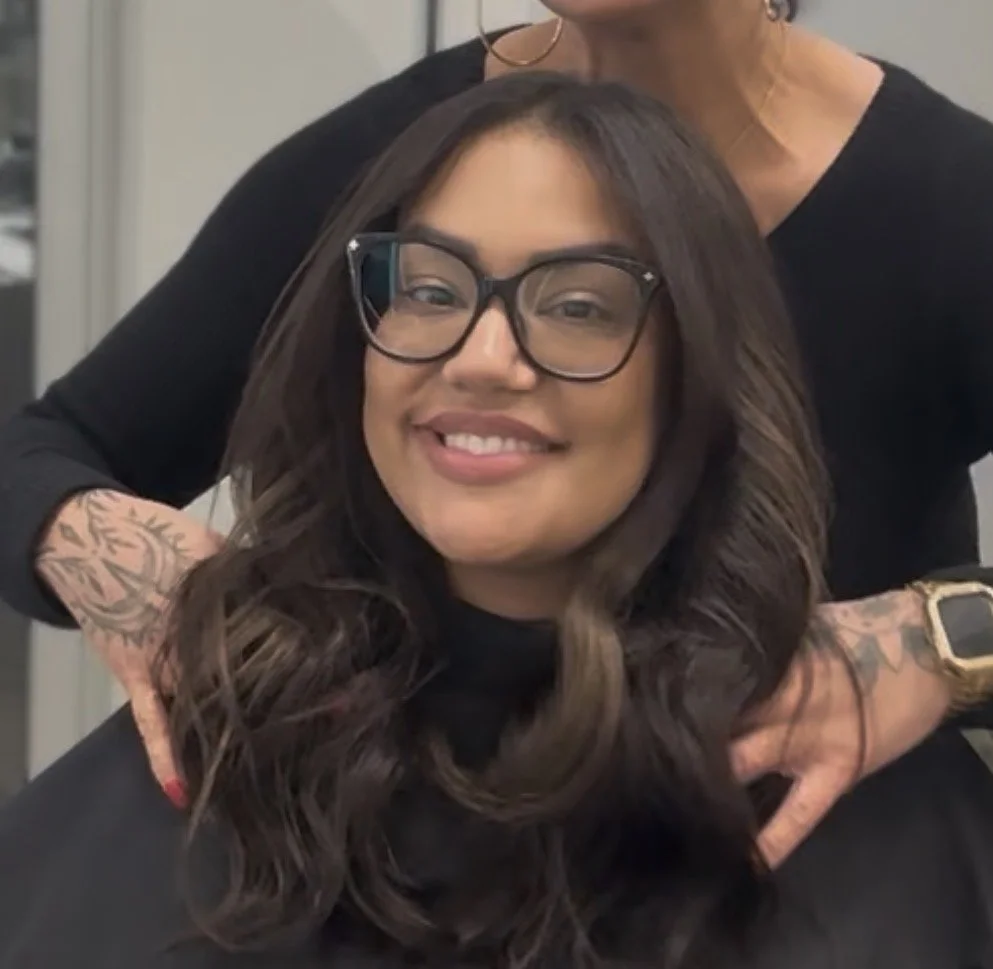 A woman with long, wavy dark hair and glasses smiling sitting in a salon chair, with a hairstylist standing behind her.