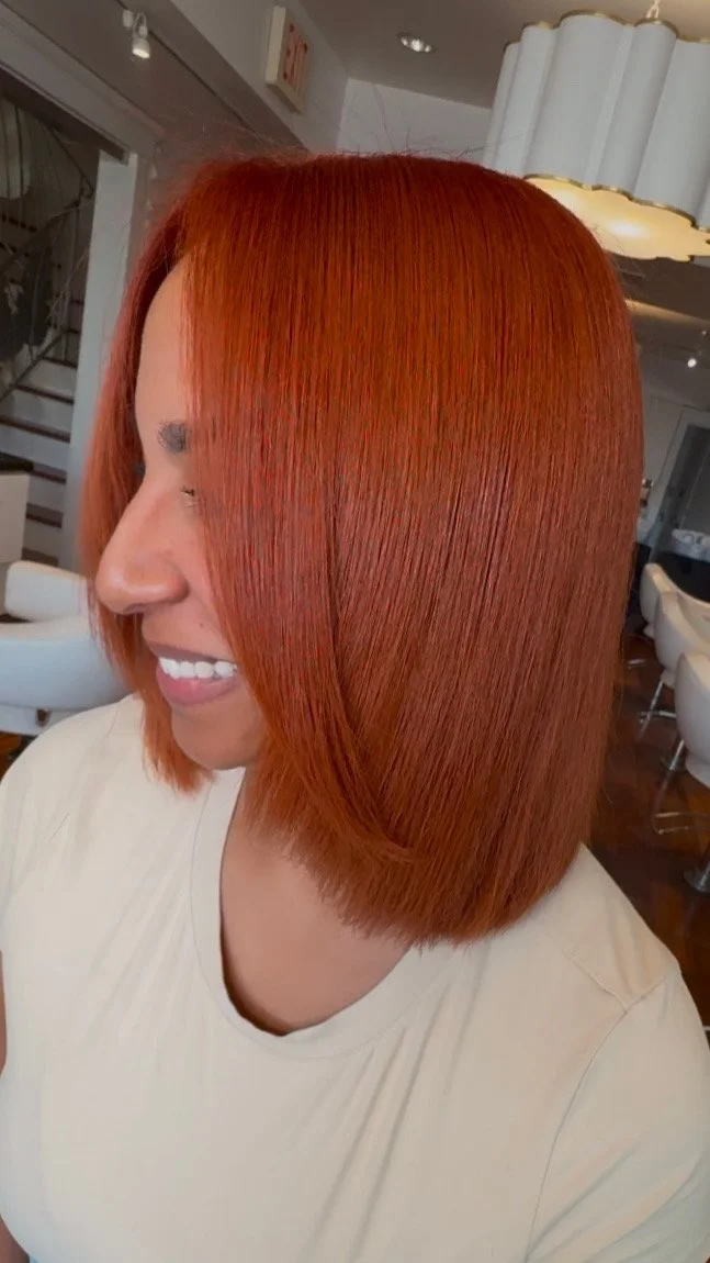 Woman with vibrant red, straight, shoulder-length hair smiling inside a modern salon.