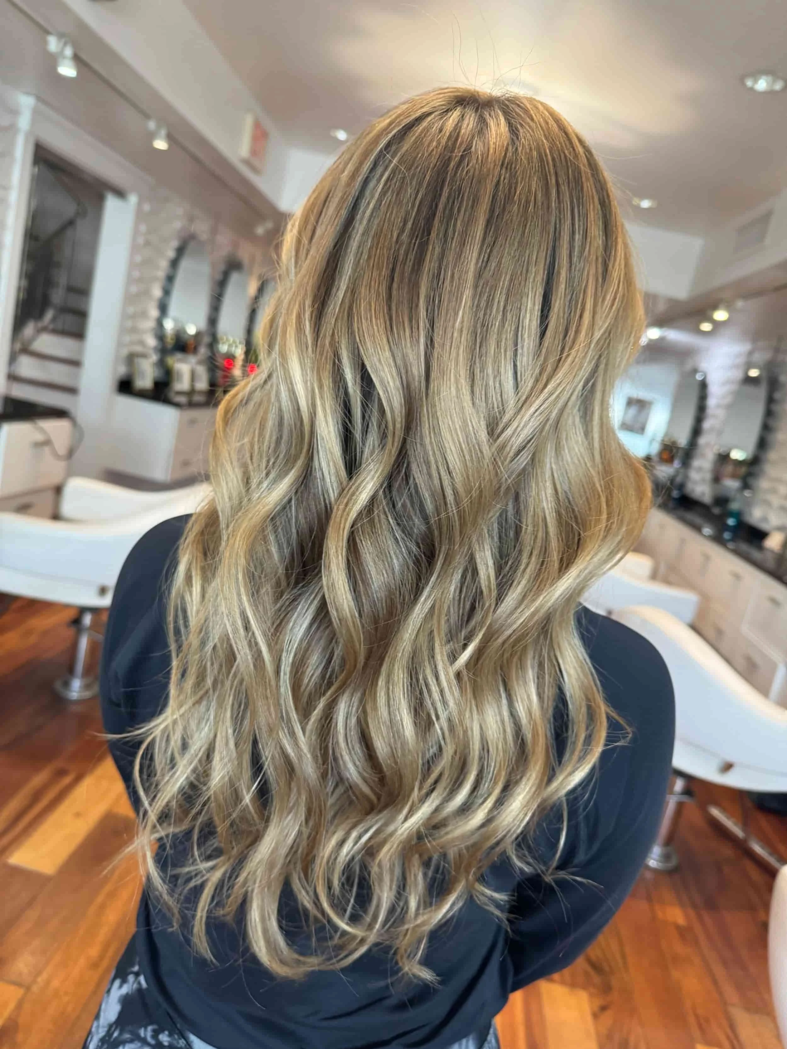 Back view of a woman with long, wavy blonde hair in a salon.