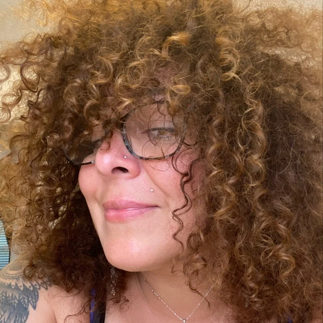 A woman with curly red hair, wearing glasses, smiling with her eyes closed, showing her nose piercings and shoulder tattoo.