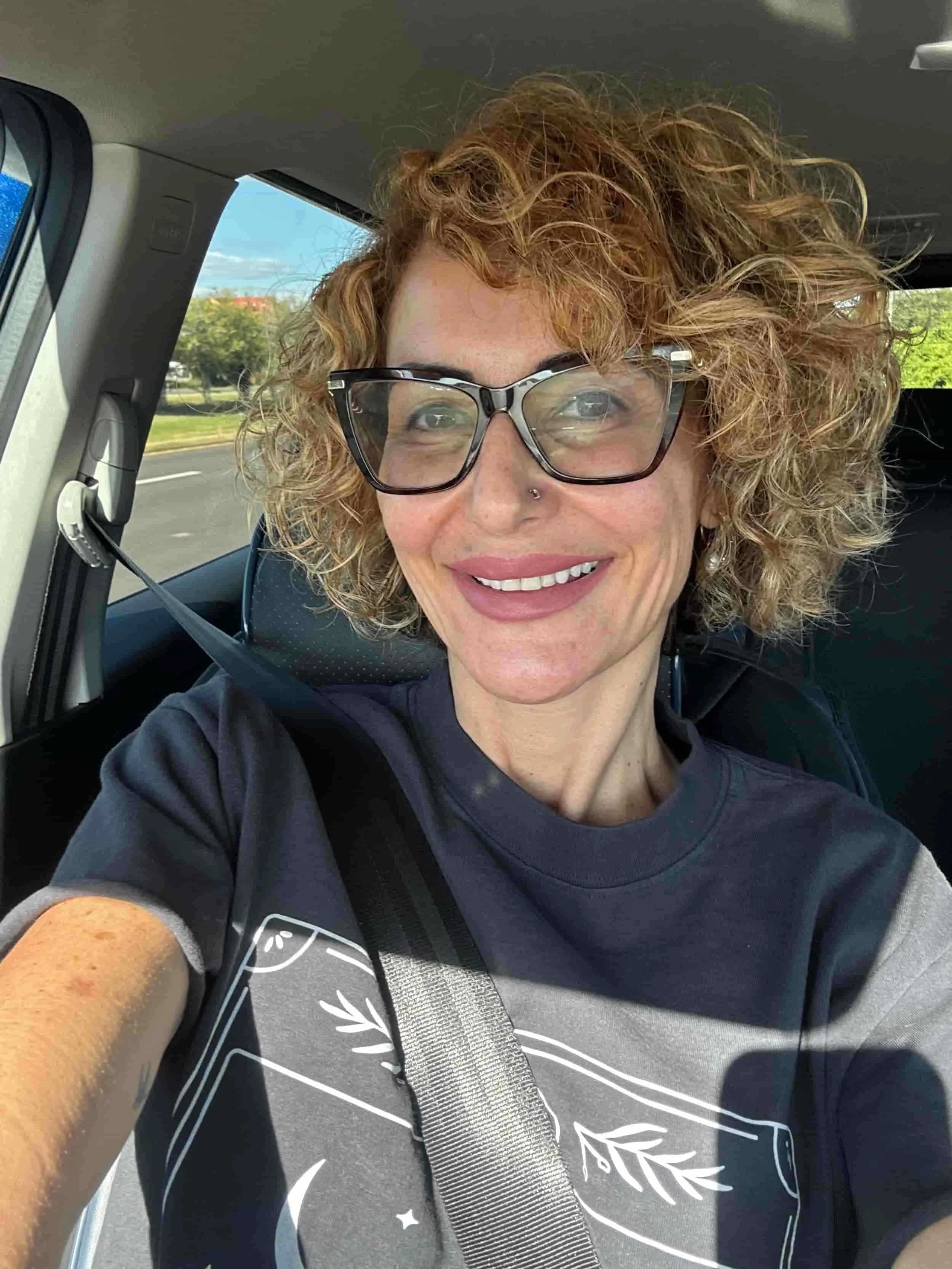 A woman with curly blonde hair, glasses, and a nose piercing, smiling in a car, wearing a dark t-shirt with a graphic design, and a seatbelt.