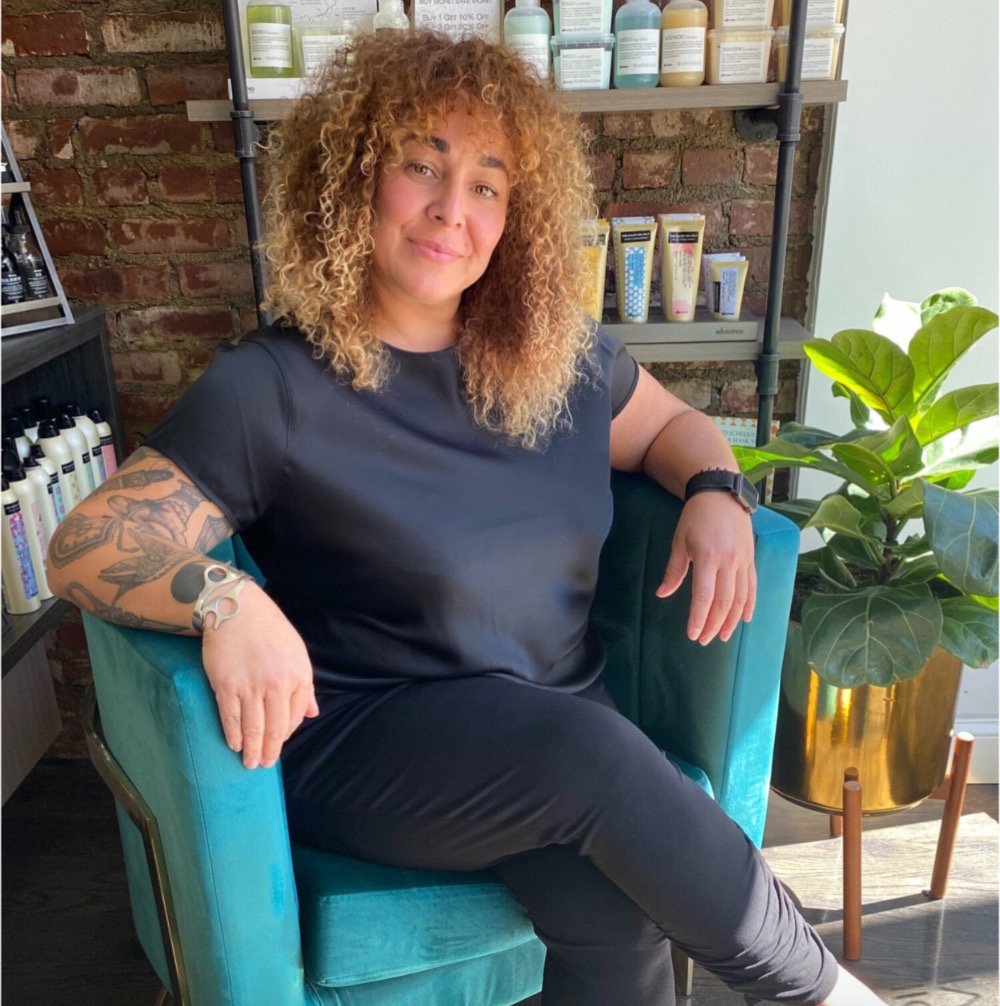 Woman sitting in a teal armchair with a brick wall and shelves with bottles in the background. She has curly hair, tattoos on her left arm, and is wearing a black shirt and black pants.