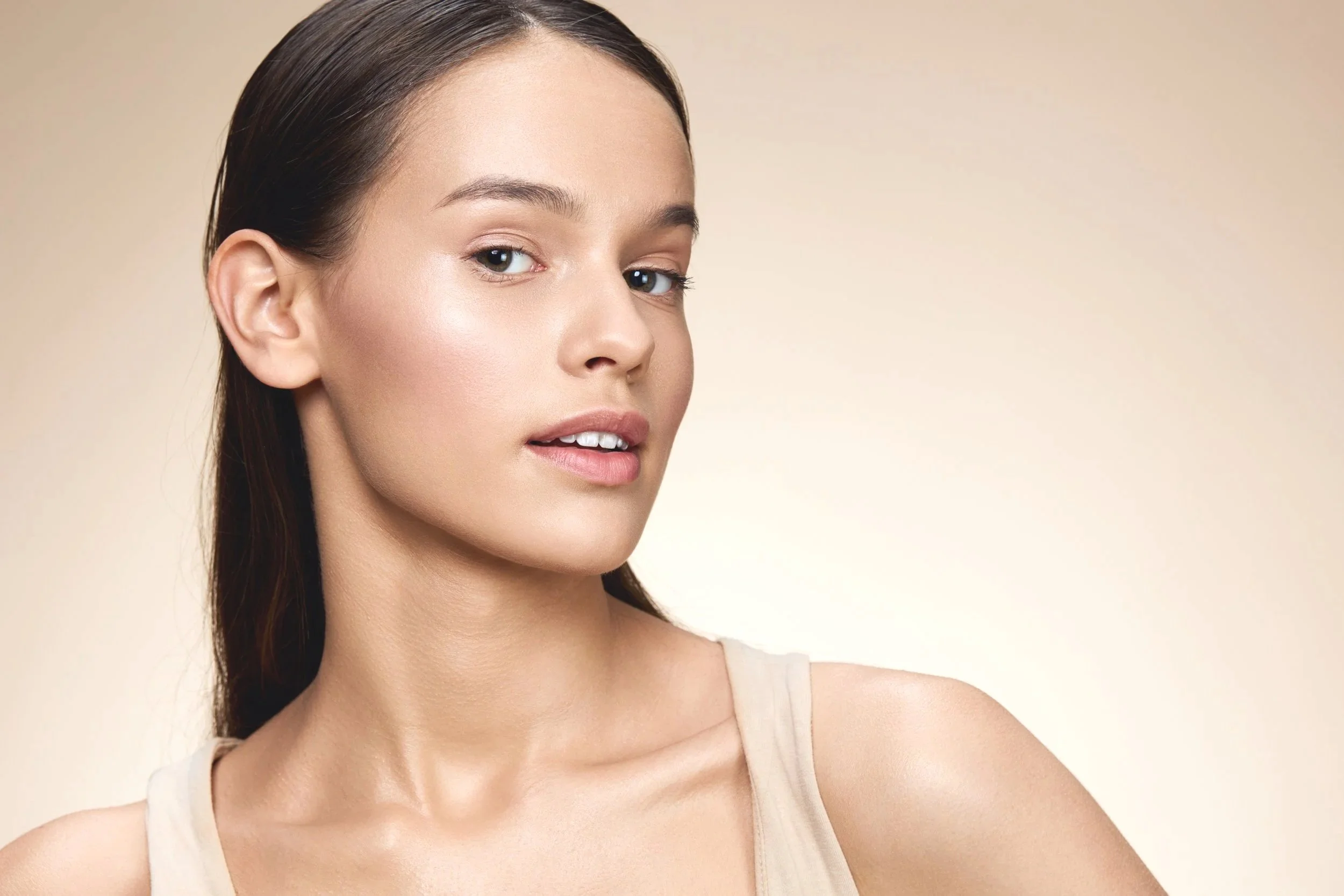 Close-up of a young woman with fair skin, brown hair, and natural makeup, looking slightly to the side against a beige background.