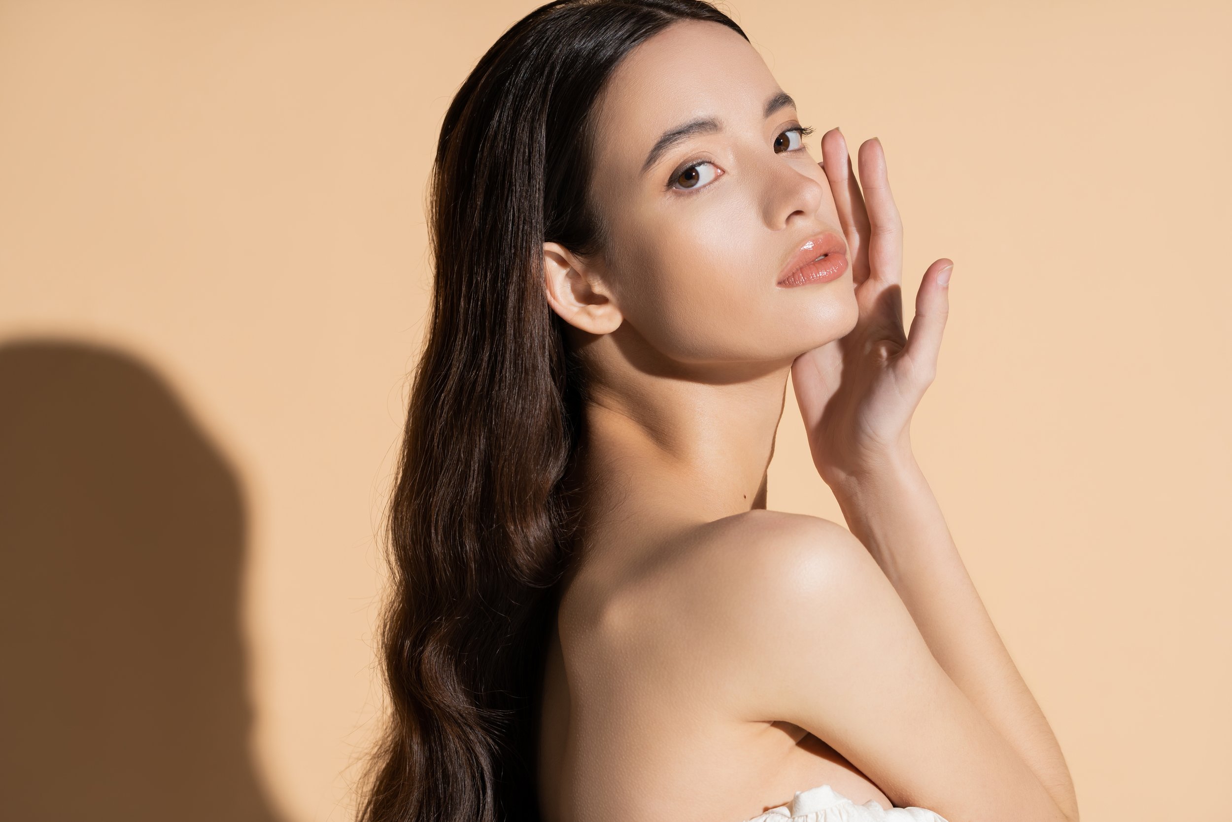 A woman with long brown hair, natural makeup, and glossy lips posing against a beige background.