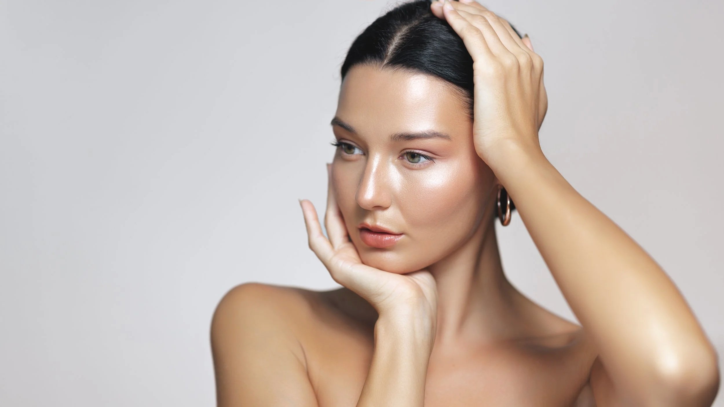 Close-up of a woman with her hand on her head and chin, looking thoughtfully to the side, with moisturized skin and dark hair.