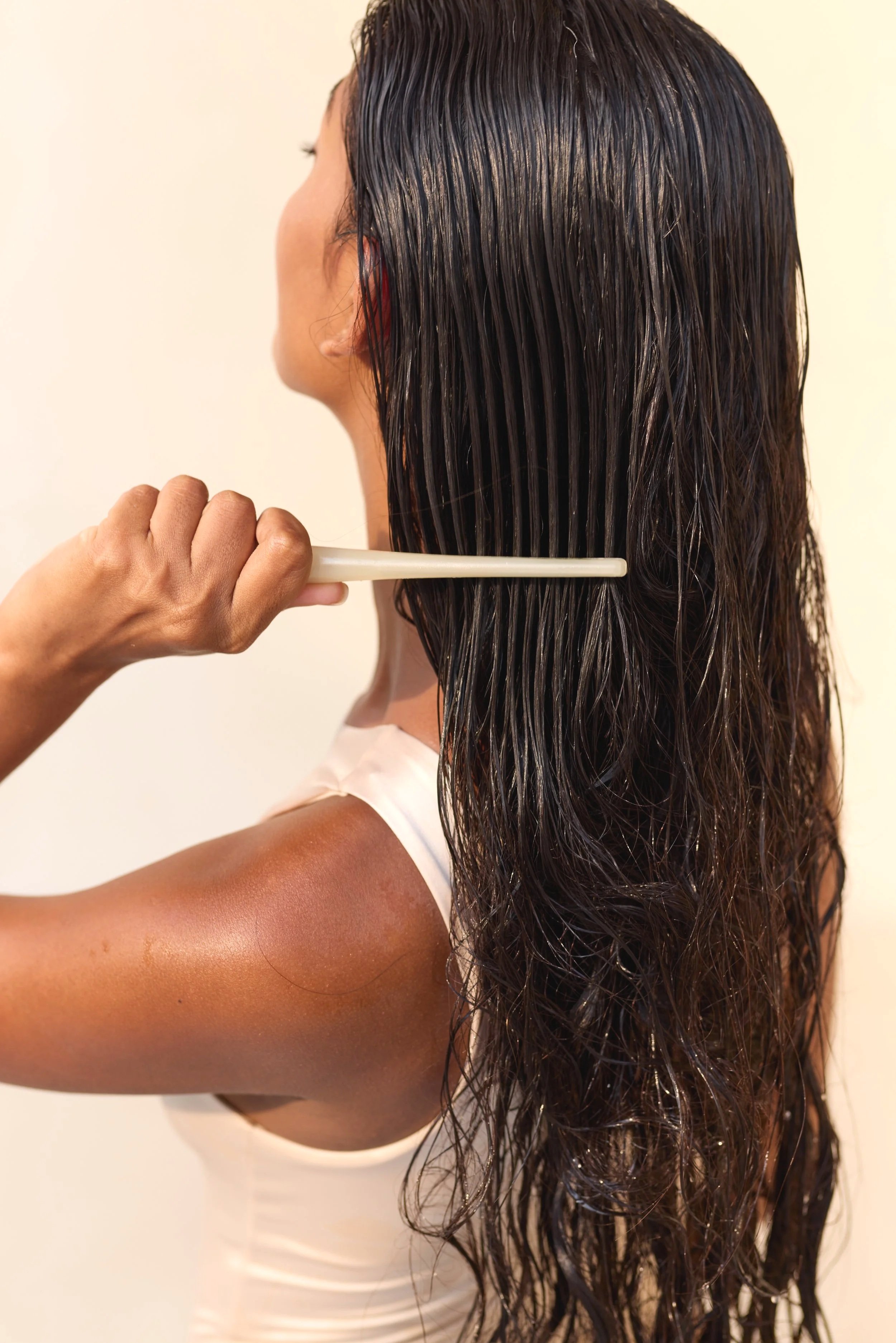A woman with wet, dark hair combs her hair with a white wide-tooth comb, focusing on the back of her head and shoulder.