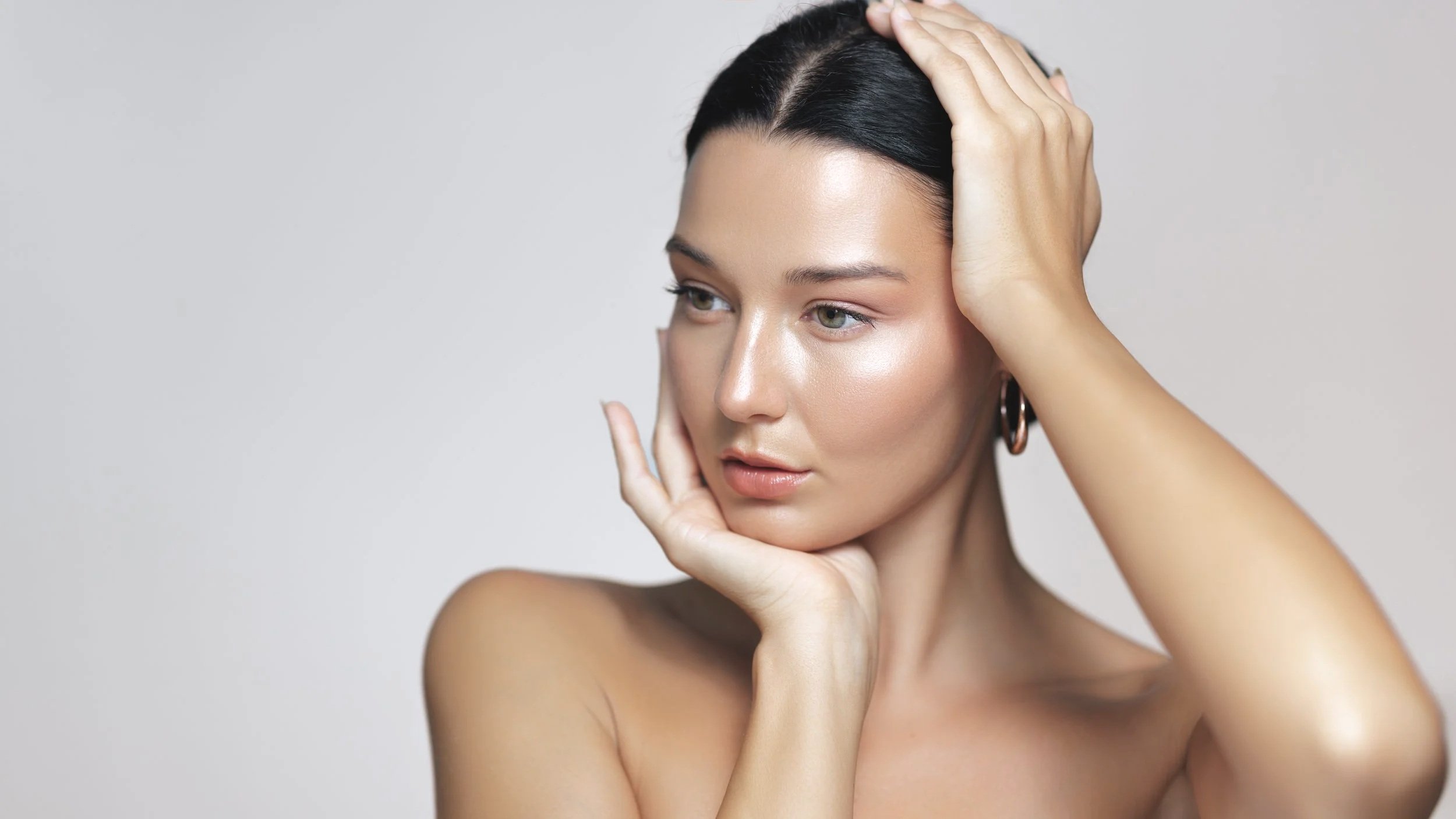 A woman with fair skin and dark hair posing against a plain light gray background, with one hand touching her face and the other on her head, gazing pensively.