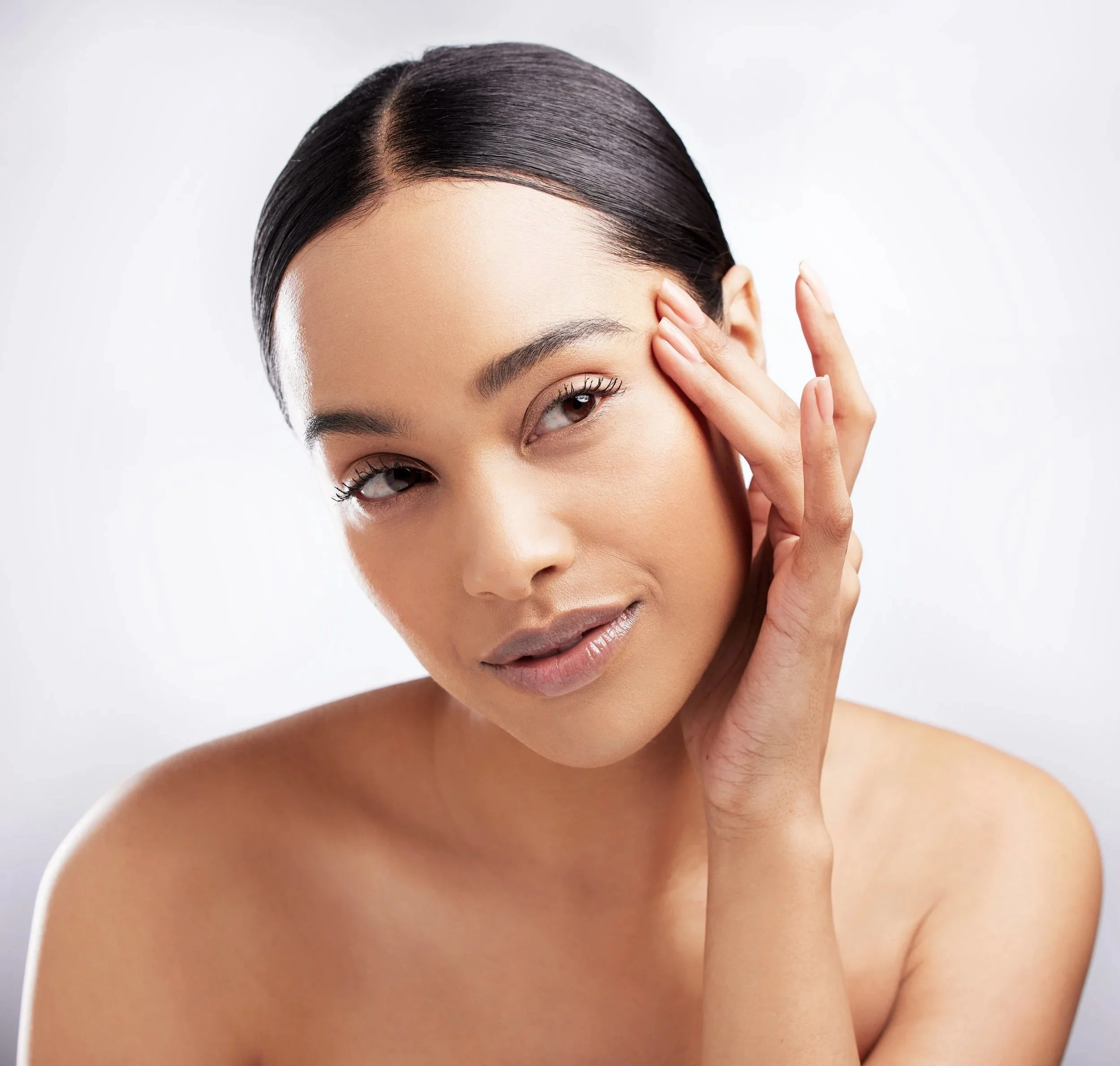 A close-up of a young woman with dark hair styled in a sleek bun touching her face with one hand, looking slightly to the side, against a plain light background.