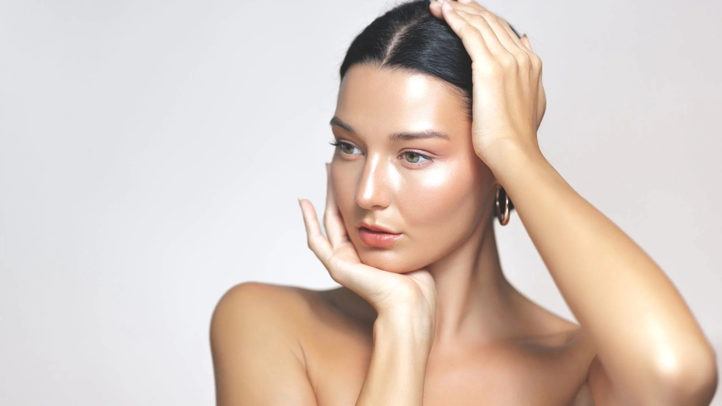 A young woman with dark hair and hoop earrings gently touches her face and head, looking contemplative against a plain light background.