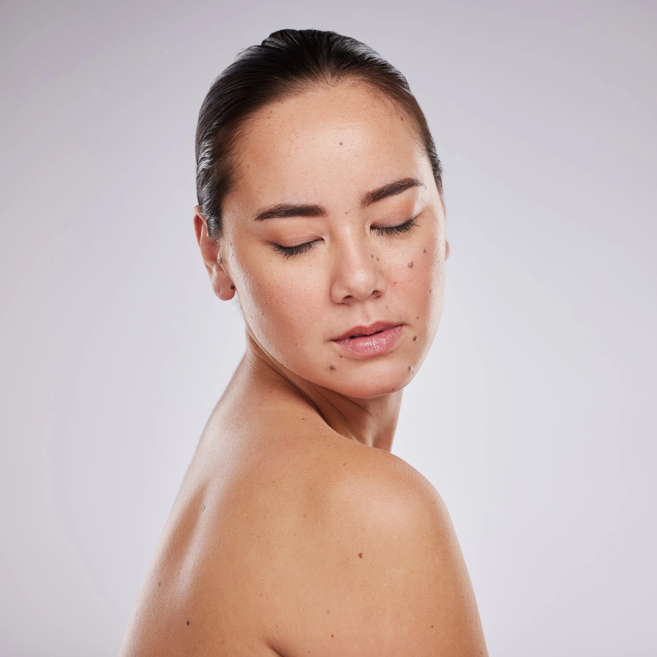 Close-up of a woman with her eyes closed, showing her shoulder and face with moles and freckles, against a plain gray background.