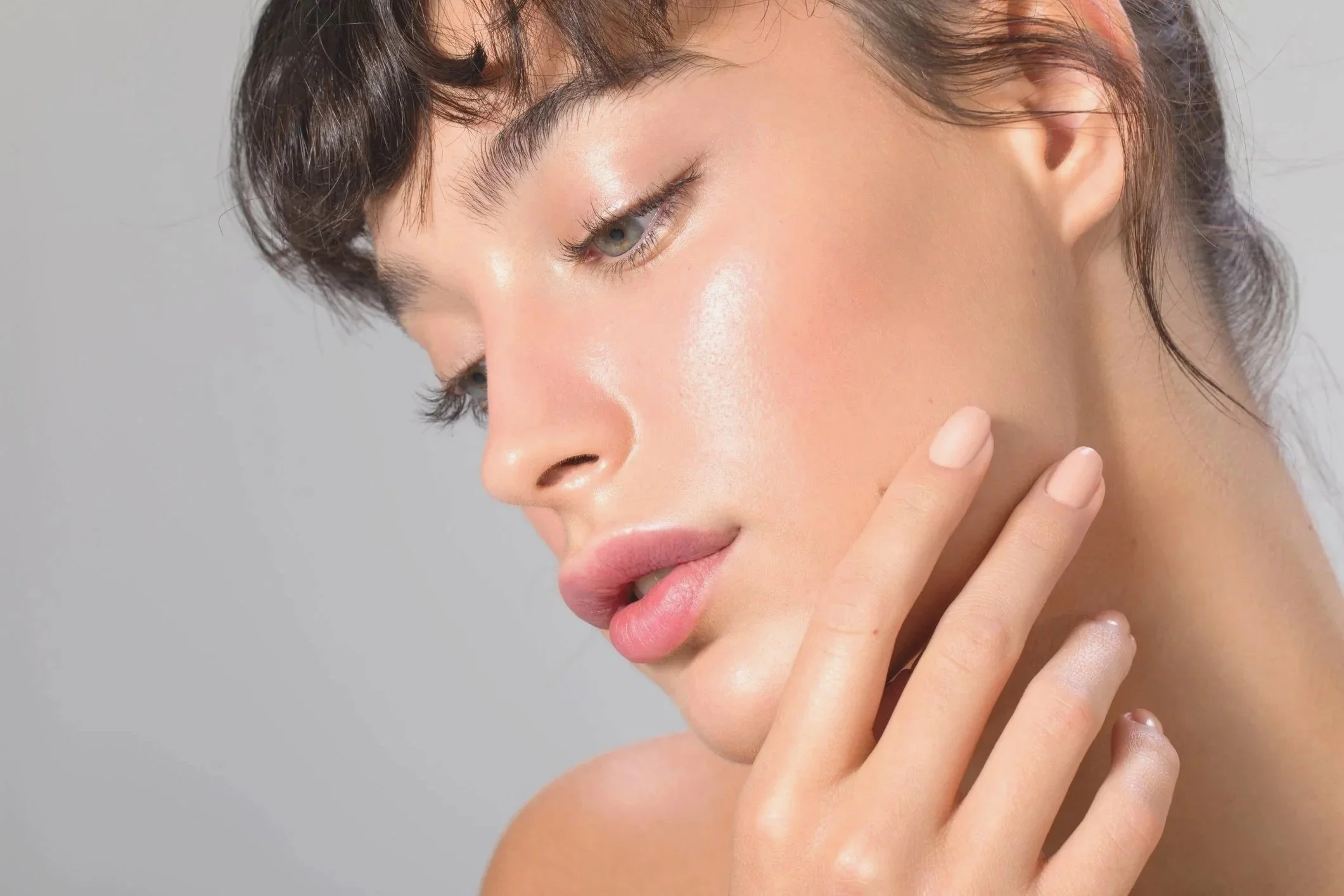 Close-up of a woman's face with smooth skin, gently touching her cheek with her fingers, against a neutral background.