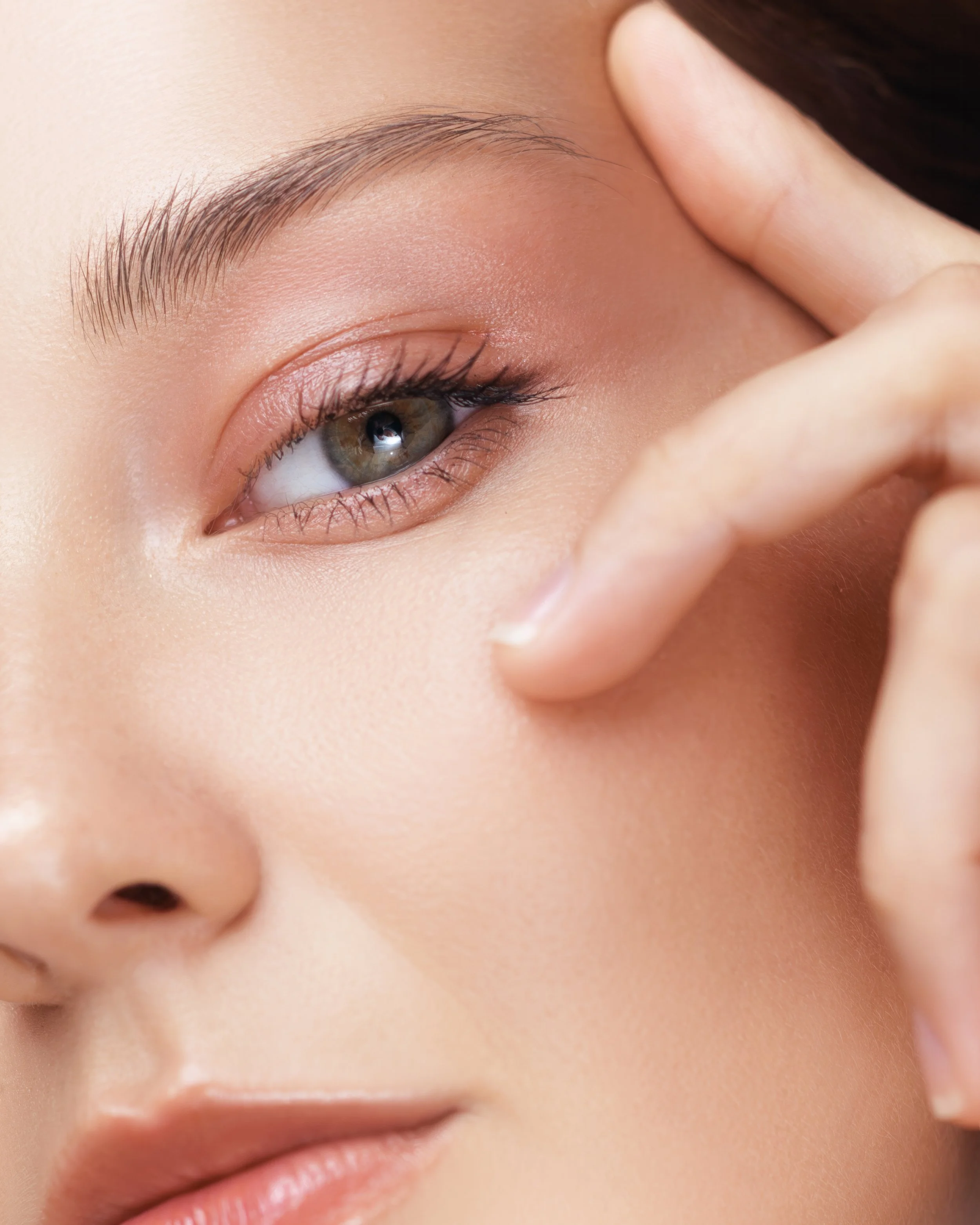 Close-up of a woman's face showcasing clear skin, with her eye partially closed and her finger touching her temple.