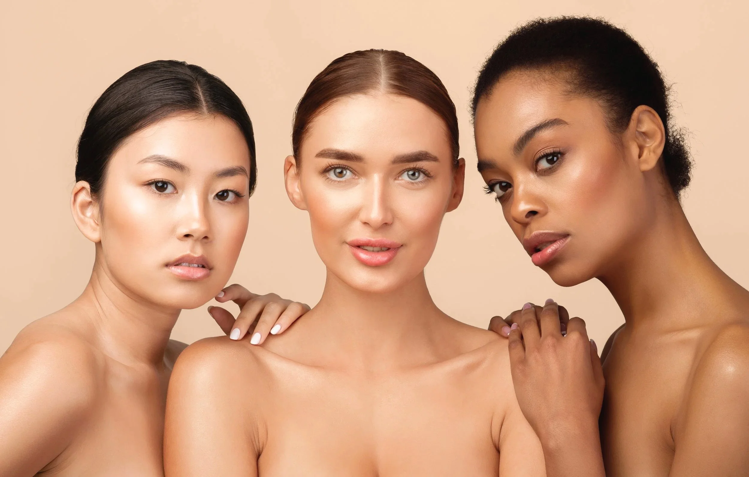 Three women of diverse ethnicities with natural makeup and bare shoulders posing together against a beige background.