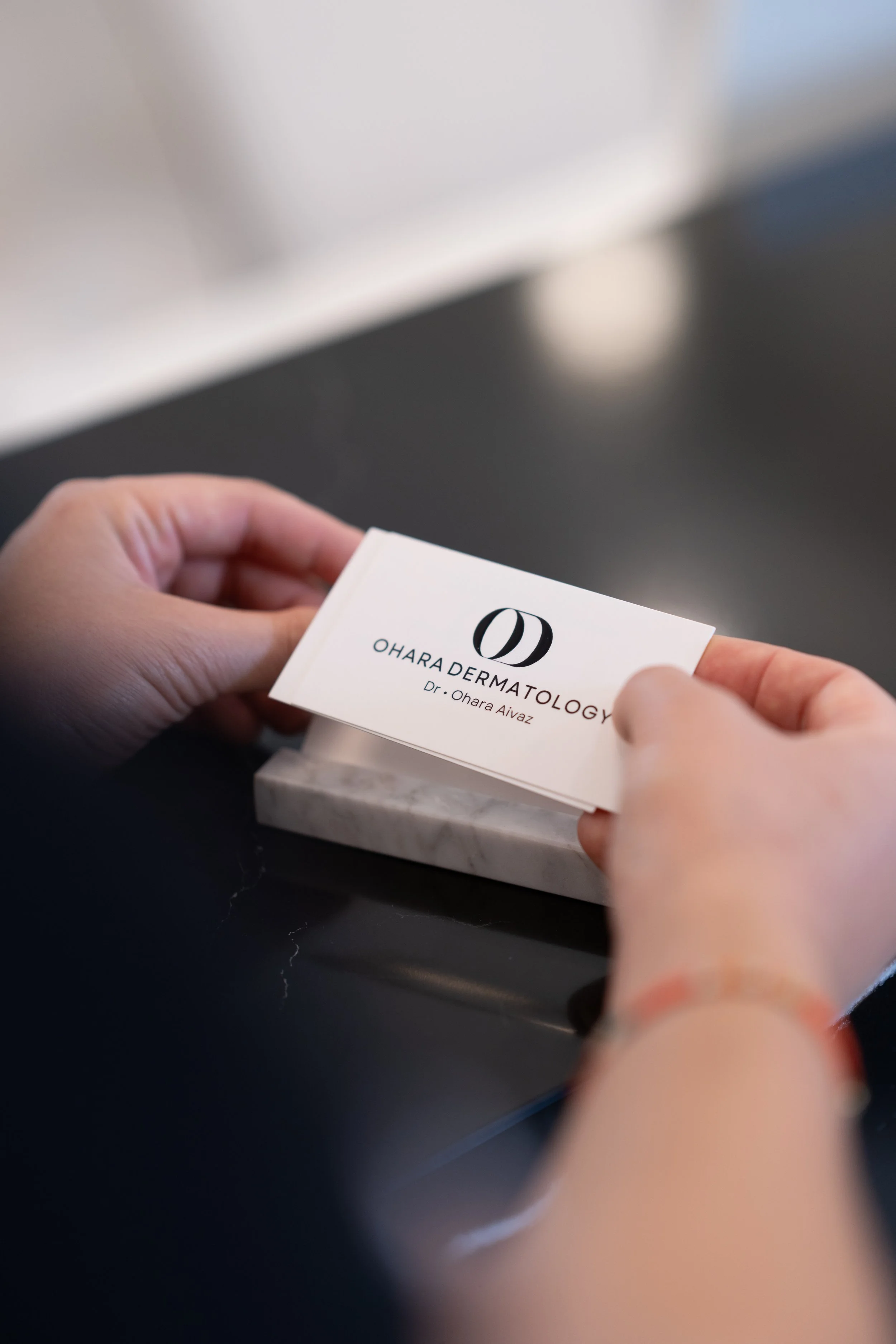 Close-up of a person holding a business card for Ohara Dermatology with the name Dr. Ohara Aivaz, resting on a marble desk.