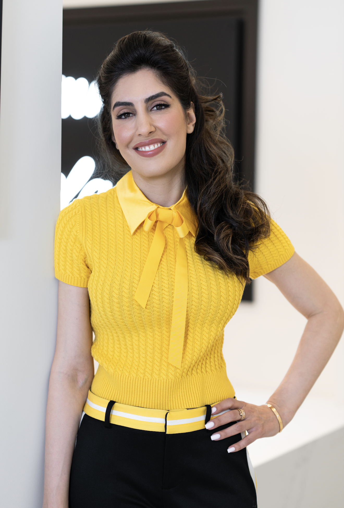 A woman with long wavy dark hair, smiling, wearing a yellow knit top with short sleeves, a yellow collar with a tie, and black pants with a yellow and white striped belt, standing indoors next to a white wall.