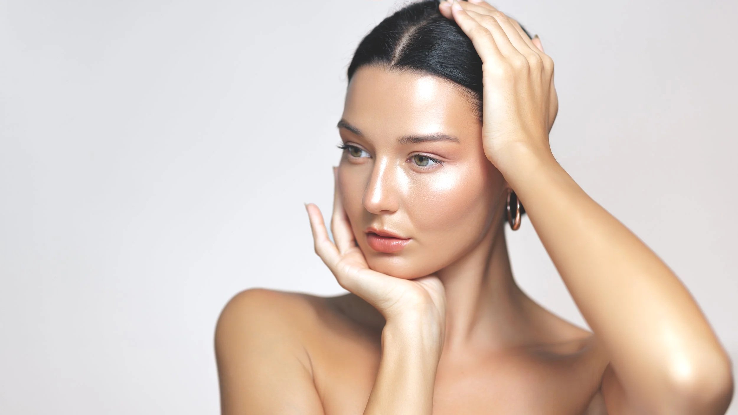 A young woman with black hair, minimal makeup, and earrings, holding her face with one hand and her head with the other, looking to the side with a neutral expression against a plain background.