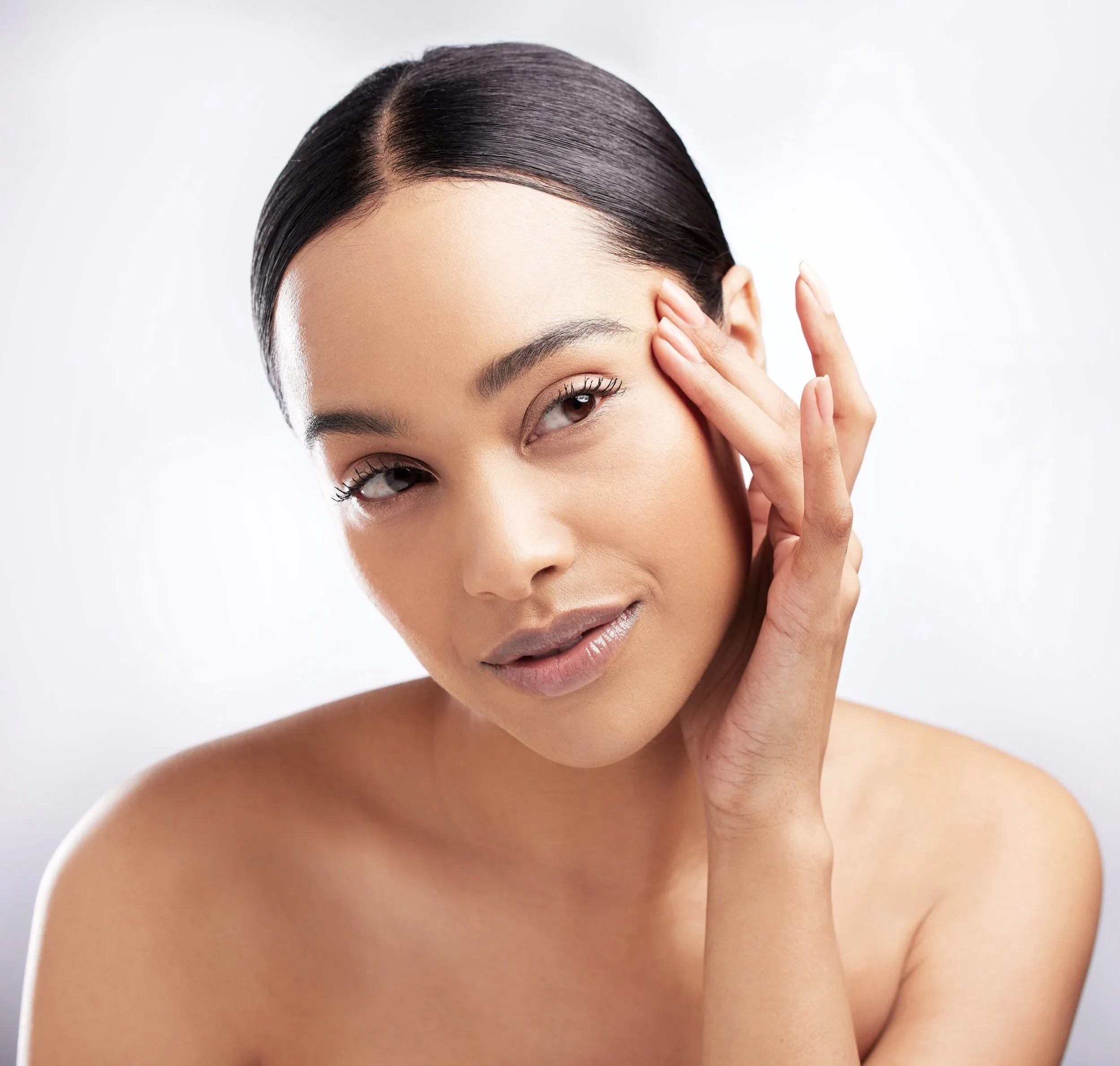 A woman with smooth, dark hair touching her face delicately, showcasing clear skin and minimal makeup against a plain white background.